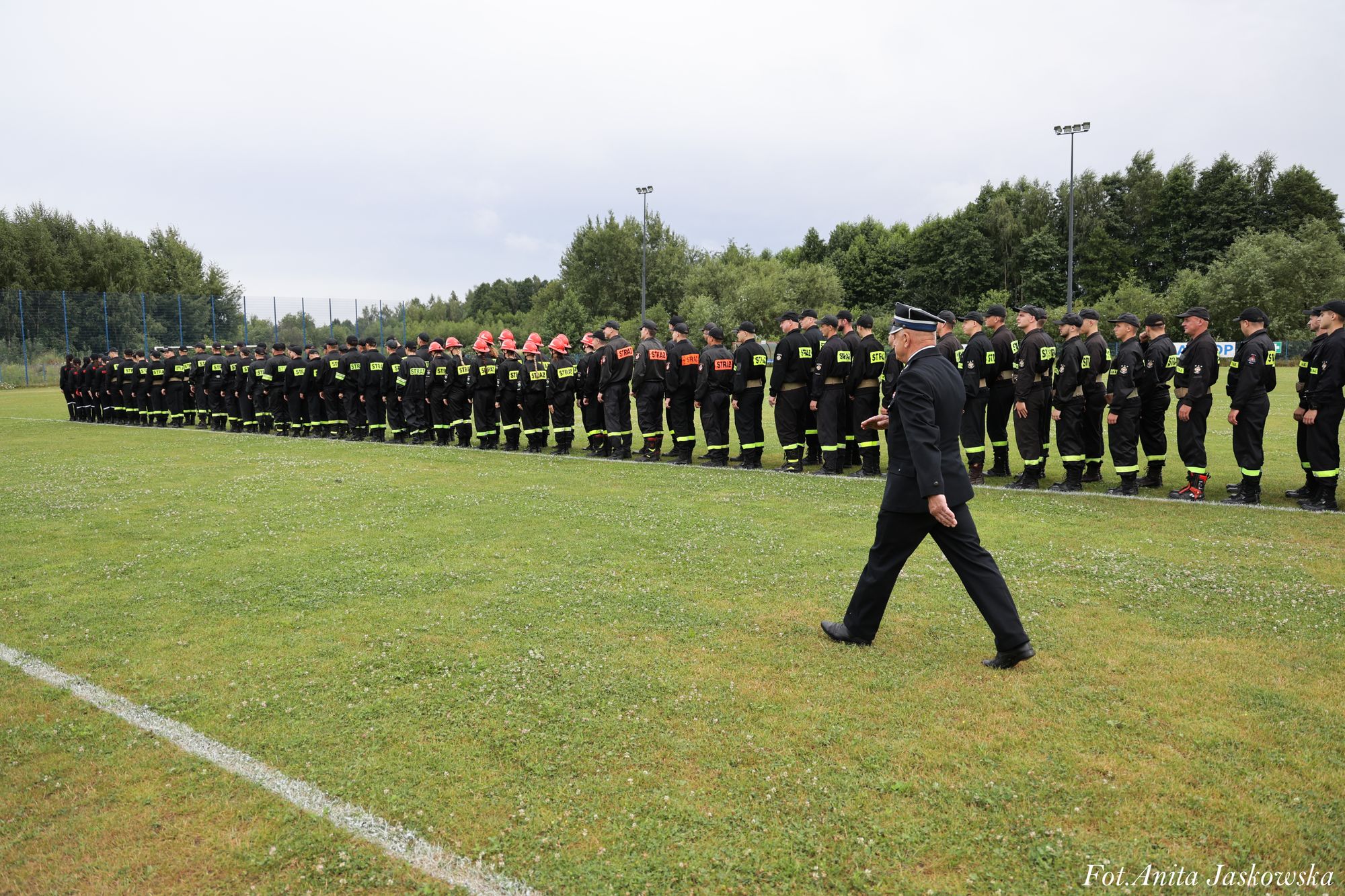Szereg osób w czarnych mundurach z żółtymi i czerwonymi napisami "STRAŻ" stoi na zielonym boisku, mężczyzna w mundurze idzie obok nich, w tle drzewa i pochmurne niebo.
