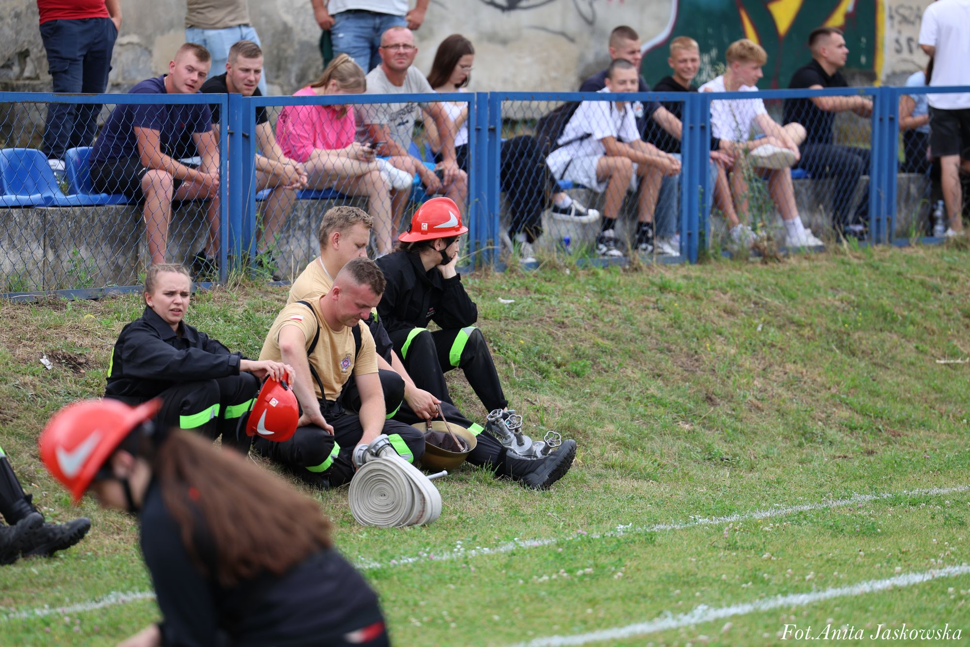 Grupa młodych strażaków w czarnych mundurach i czerwonych hełmach siedzi na trawie przy niebieskim ogrodzeniu, za którym siedzą inni ludzie na trybunach.