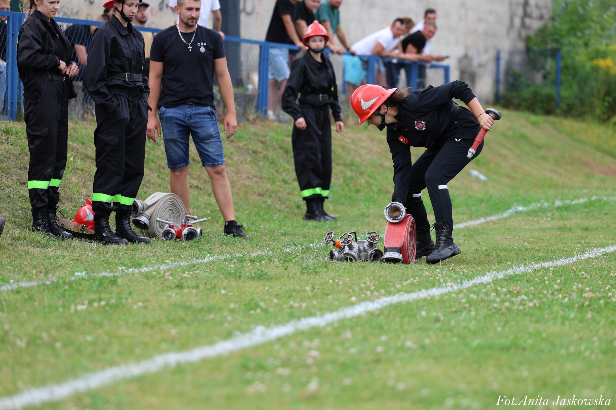 Grupa osób w czarnych ubraniach strażackich z czerwonymi hełmami, jedna osoba rozwija czerwoną strażacką linię na zielonym trawiastym boisku.