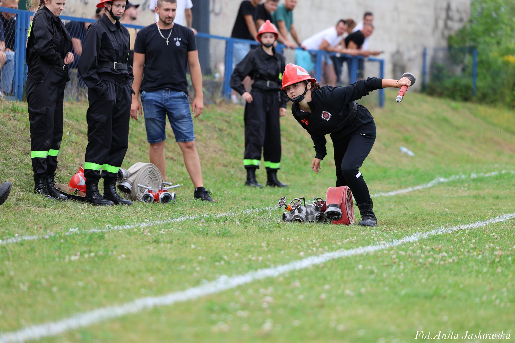 Kobieta w czarnym ubraniu i czerwonym hełmie pochyla się z wężem strażackim na zielonym boisku, obok stoi grupa osób w czarnych strojach z zielonymi pasami i mężczyzna w czarnym t-shircie i niebieskich szortach.
