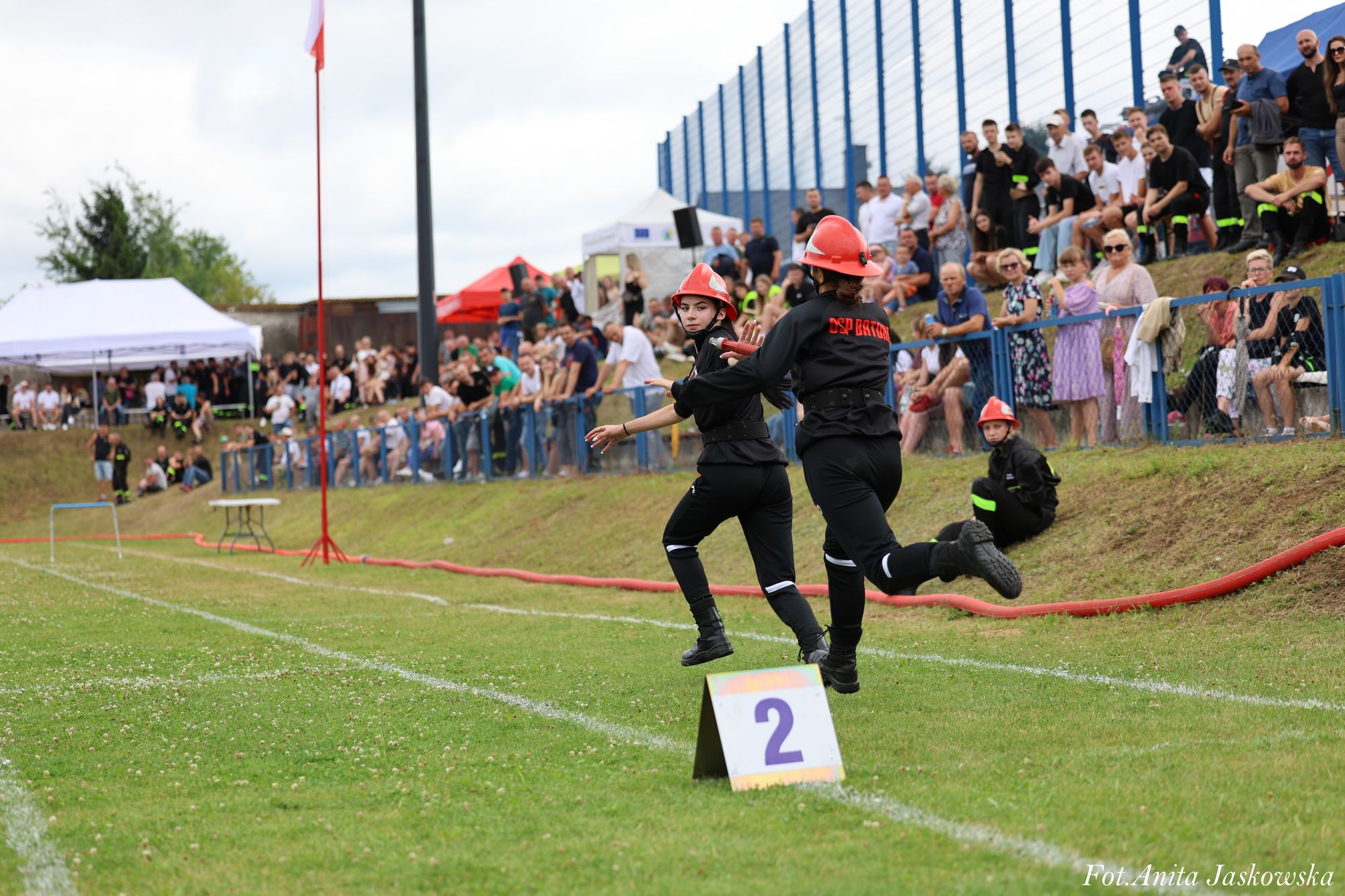 Dwie dziewczynki w czarnych strojach i czerwonych hełmach biegną obok tabliczki z numerem 2 na zielonym boisku, w tle tłum ludzi za niebieską siatką.