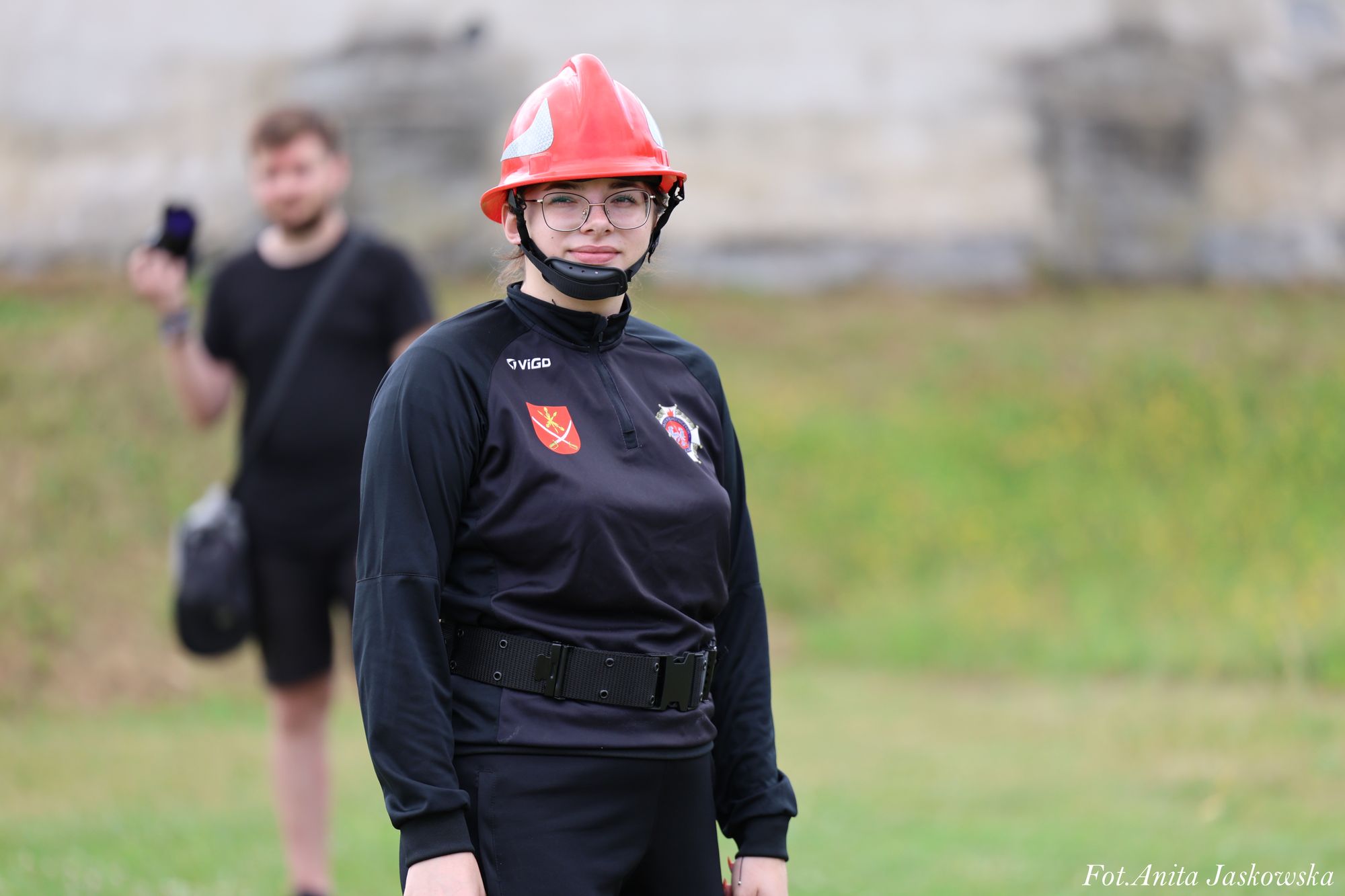 Kobieta w czarnym ubraniu z emblematami, czerwonym kaskiem i okularami stojąca na tle zieleni, w tle rozmazany mężczyzna w czarnym ubraniu.