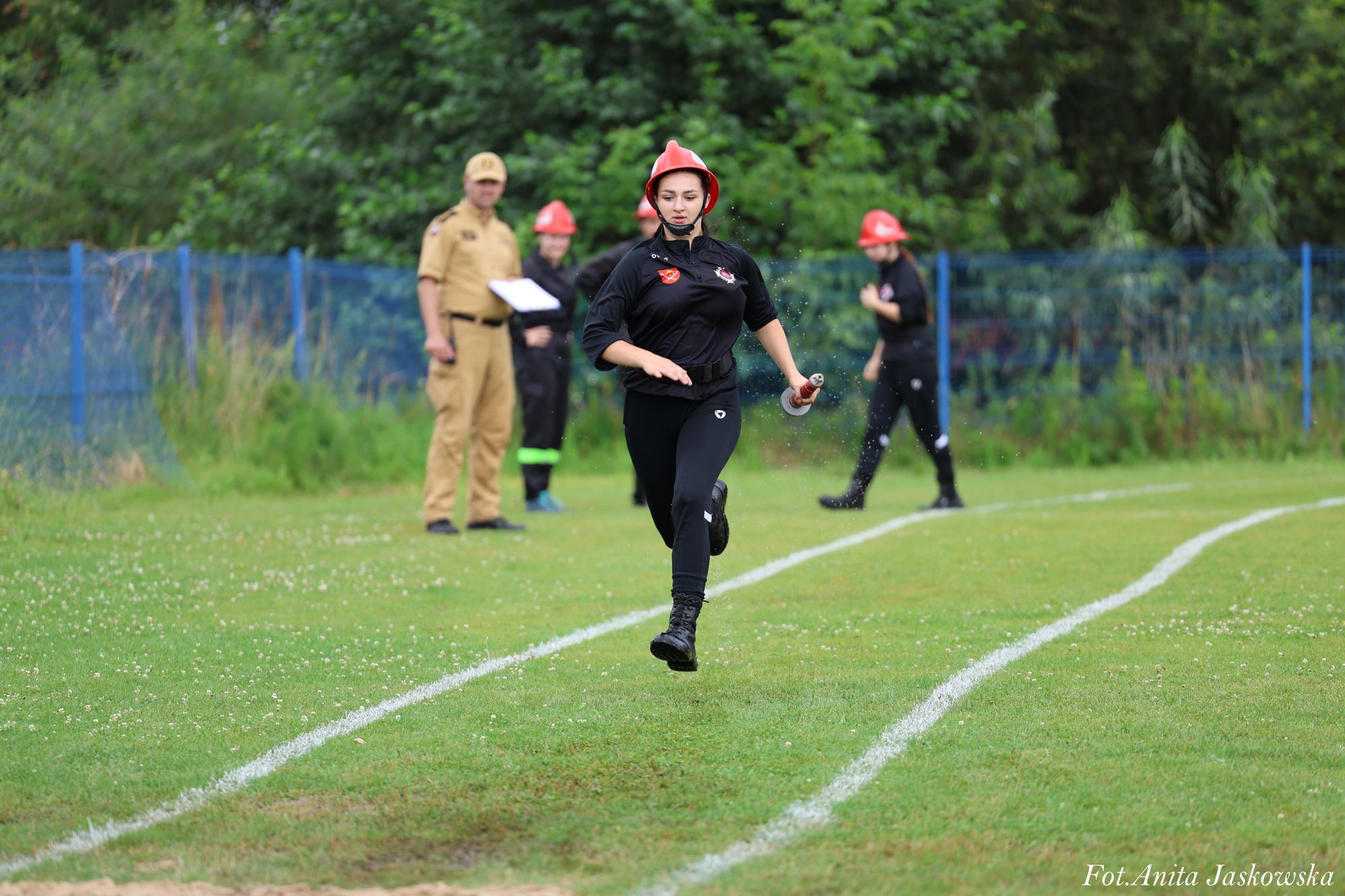 Kobieta w czarnym ubraniu i czerwonym hełmie biegnie po zielonym boisku sportowym z białymi liniami, w tle trzy osoby na tle zielonych drzew i niebieskiego ogrodzenia.