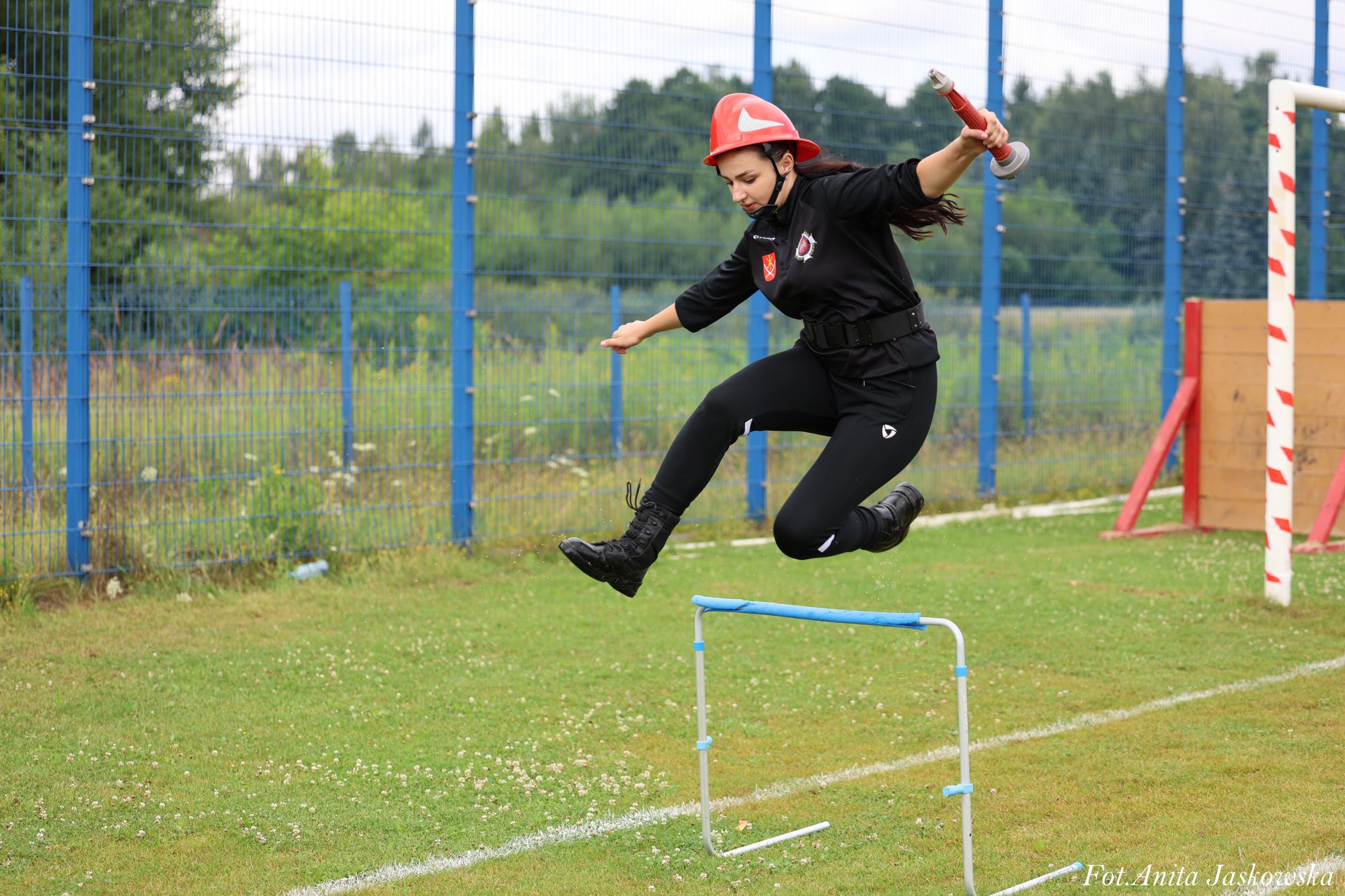 Kobieta w czarnym stroju i czerwonym hełmie skacze przez niebieską przeszkodę na zielonej trawie, trzymając czerwony wąż strażacki, w tle siatka i drzewa.
