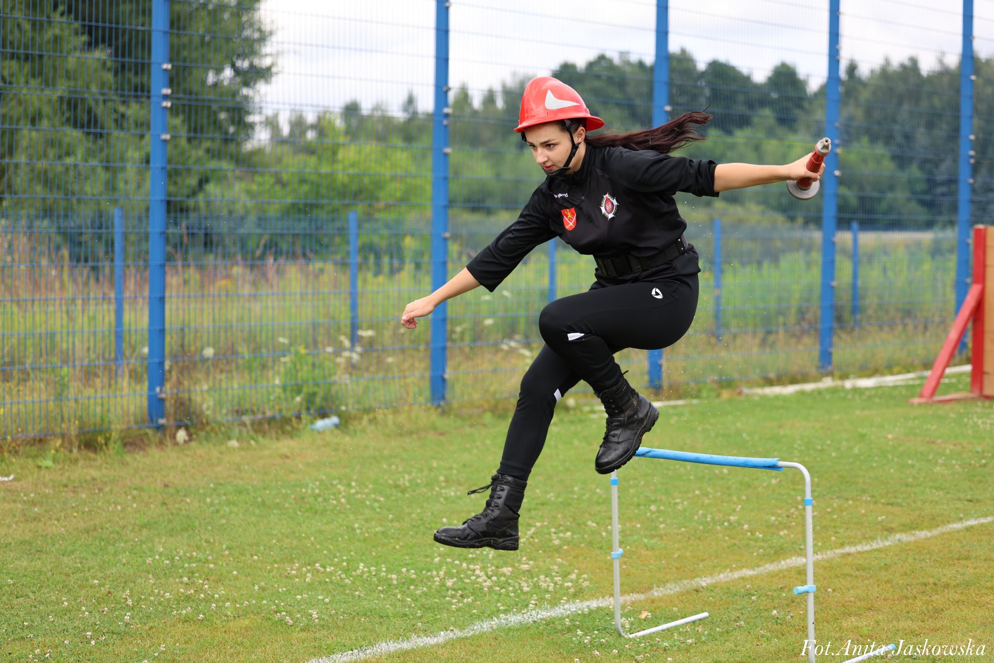Kobieta w czarnym ubraniu i czerwonym hełmie skacze przez niebieską przeszkodę na zielonym boisku z ogrodzeniem i drzewami w tle.
