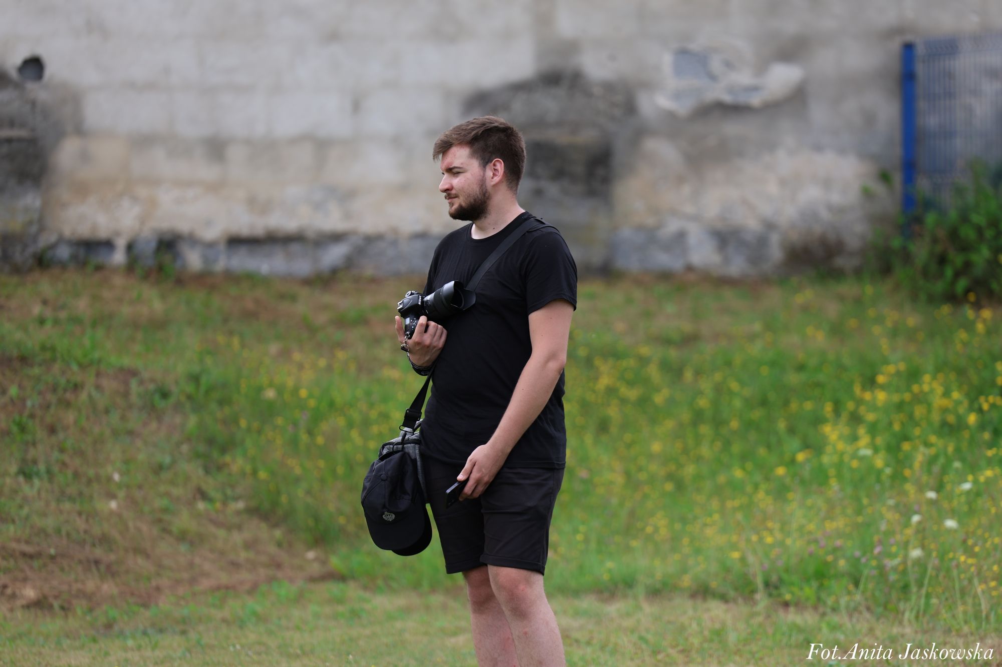 Mężczyzna w czarnym t-shircie i spodenkach stoi na trawie, trzyma aparat fotograficzny i czapkę, w tle zielone rośliny i szara, zniszczona ściana.