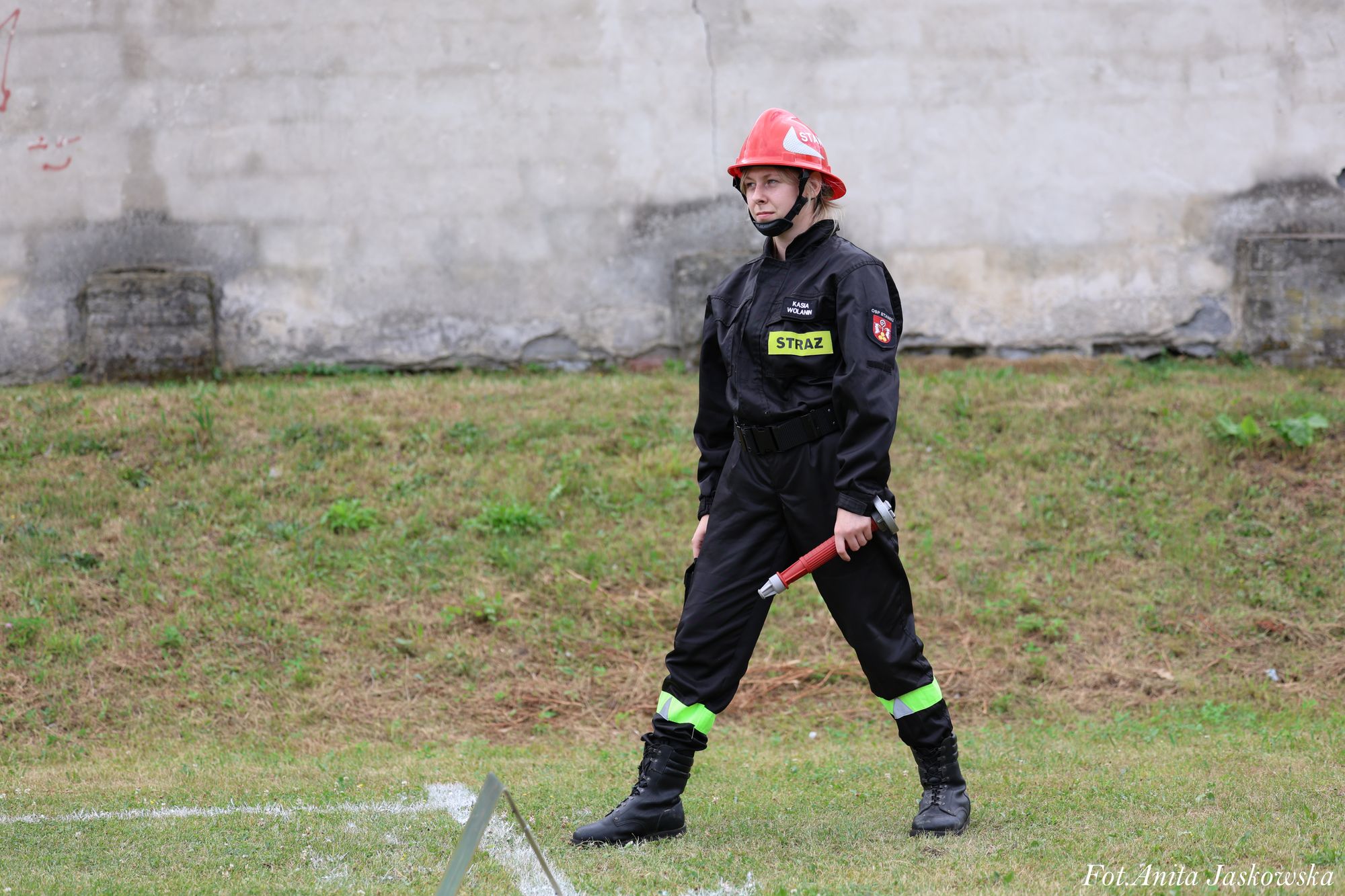 Kobieta w czarnym kombinezonie strażackim z odblaskowymi elementami i czerwonym hełmem, trzymająca czerwony sprzęt, stoi na trawie przed betonową ścianą.