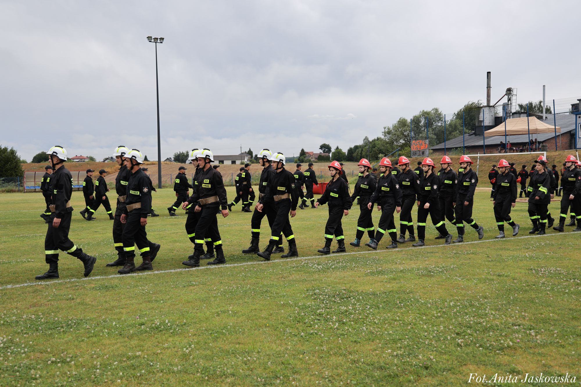 Grupa osób w mundurach strażackich z białymi i czerwonymi hełmami maszeruje po zielonym boisku sportowym pod pochmurnym niebem.