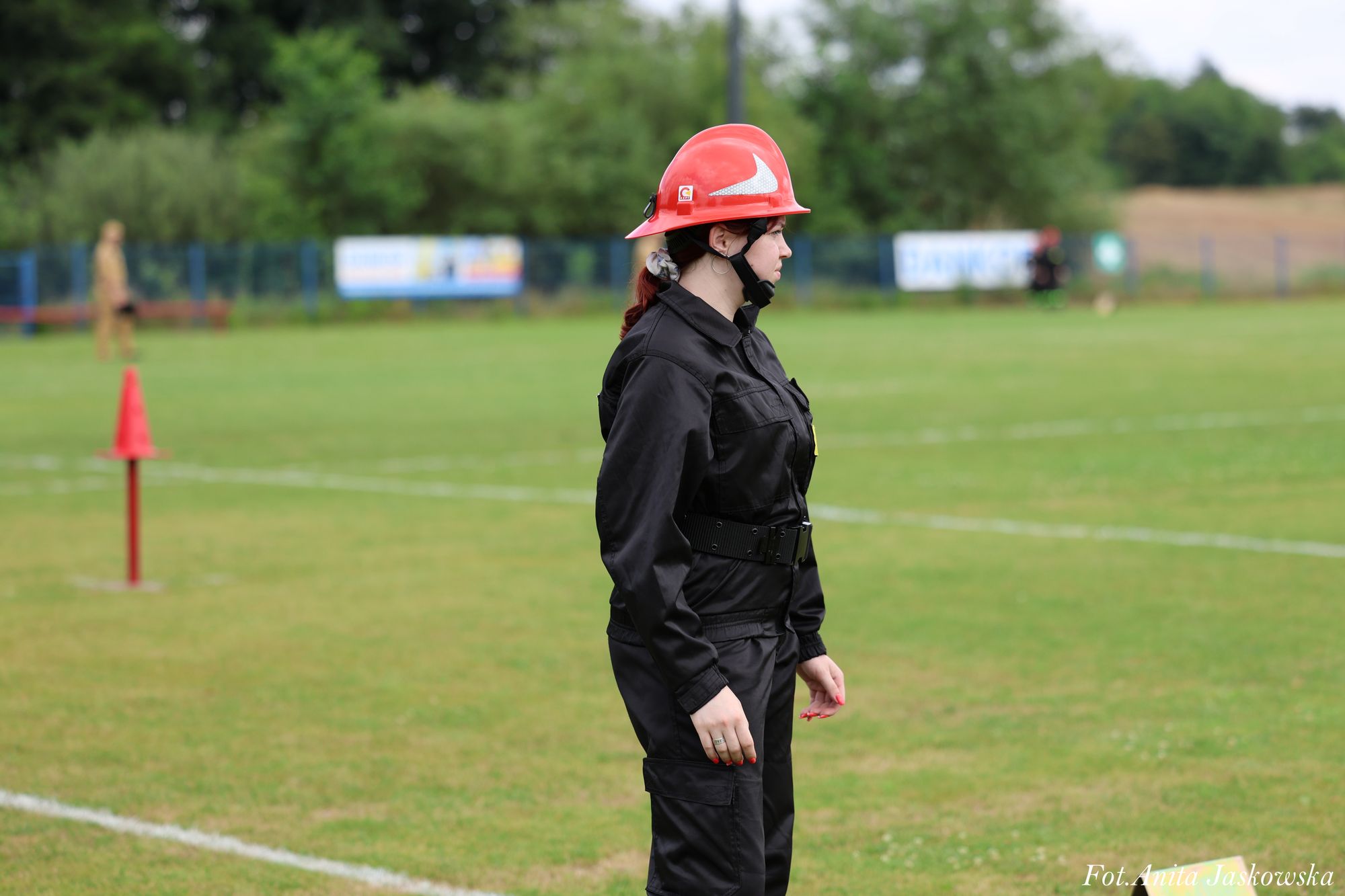Kobieta w czarnym kombinezonie i czerwonym hełmie stoi na zielonym boisku, w tle drzewa i rozmyte sylwetki osób oraz czerwony pachołek.
