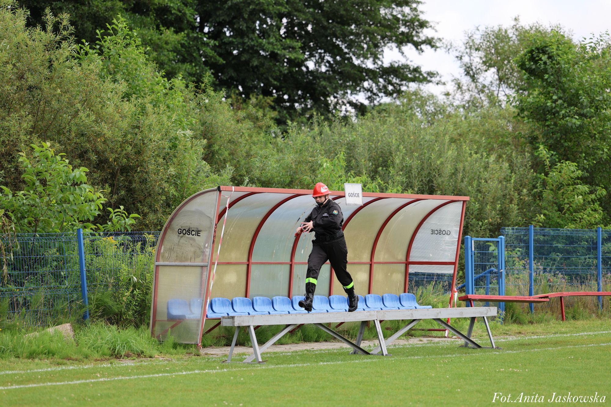 Osoba w czarnym ubraniu ochronnym i czerwonym hełmie biegnie po ławce z niebieskimi siedzeniami przed przezroczystą wiatą na zielonym boisku, w tle gęste zielone krzewy.