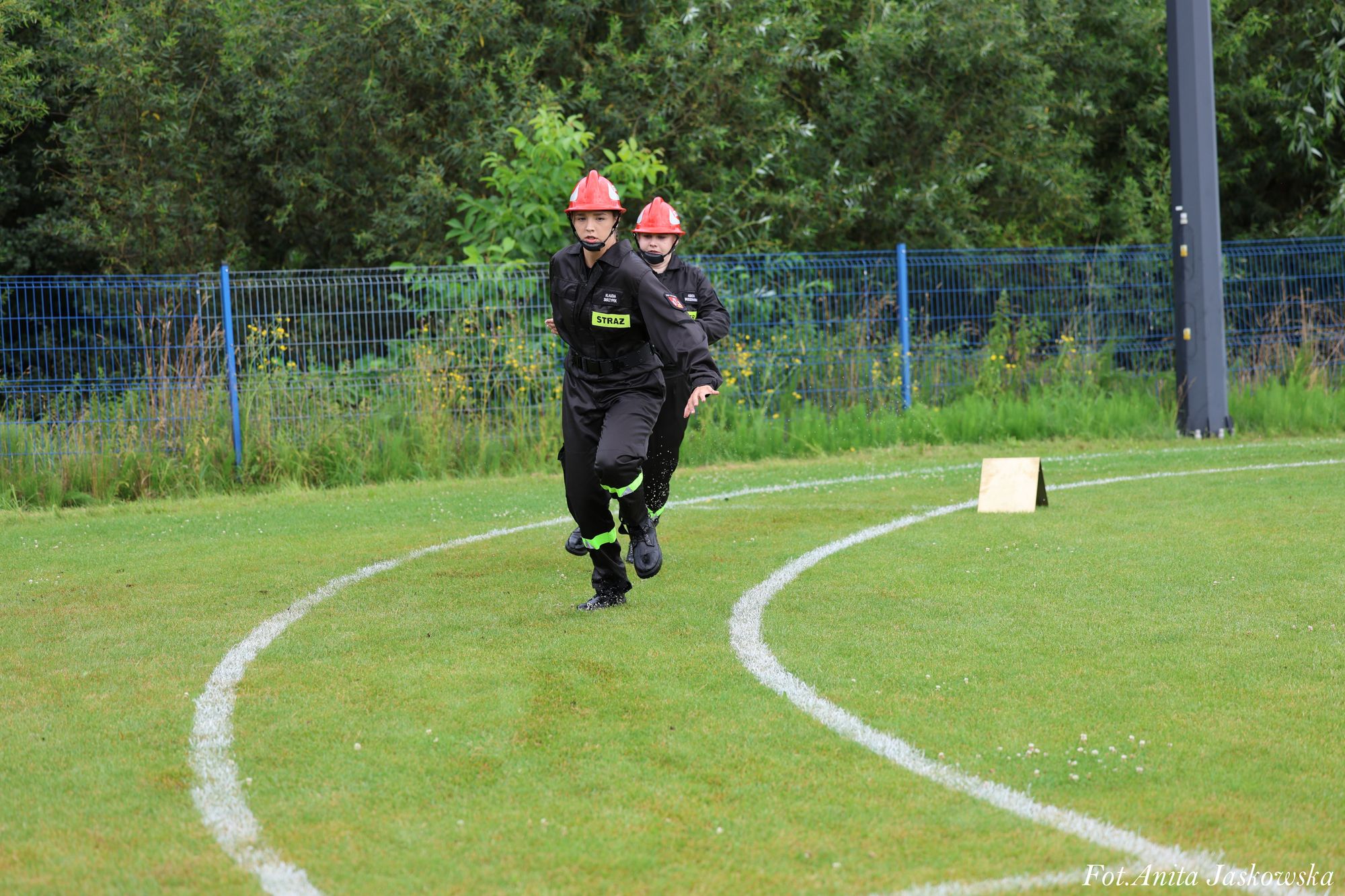 Dwóch młodych strażaków w czarnych mundurach i czerwonych hełmach biegnie po zielonej trawie z białymi liniami, w tle zielone krzewy i niebieski płot.