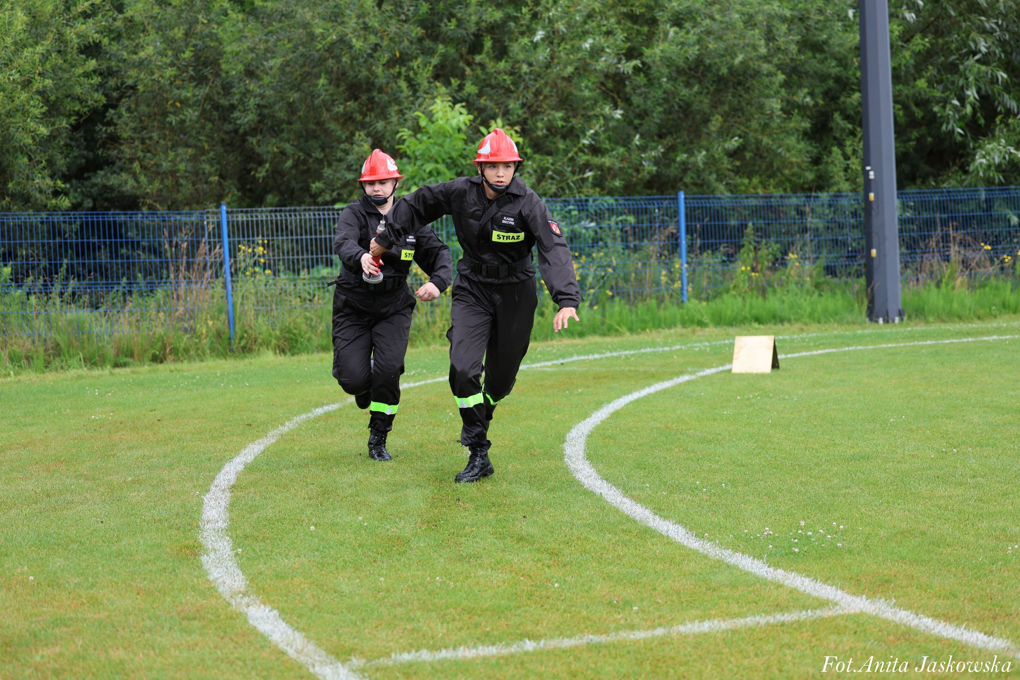 Dwie osoby w czarnych kombinezonach z napisem "STRAŻ", czerwonych kaskach i butach biegają po zielonym boisku z białymi liniami, w tle zielone krzewy i niebieskie ogrodzenie.