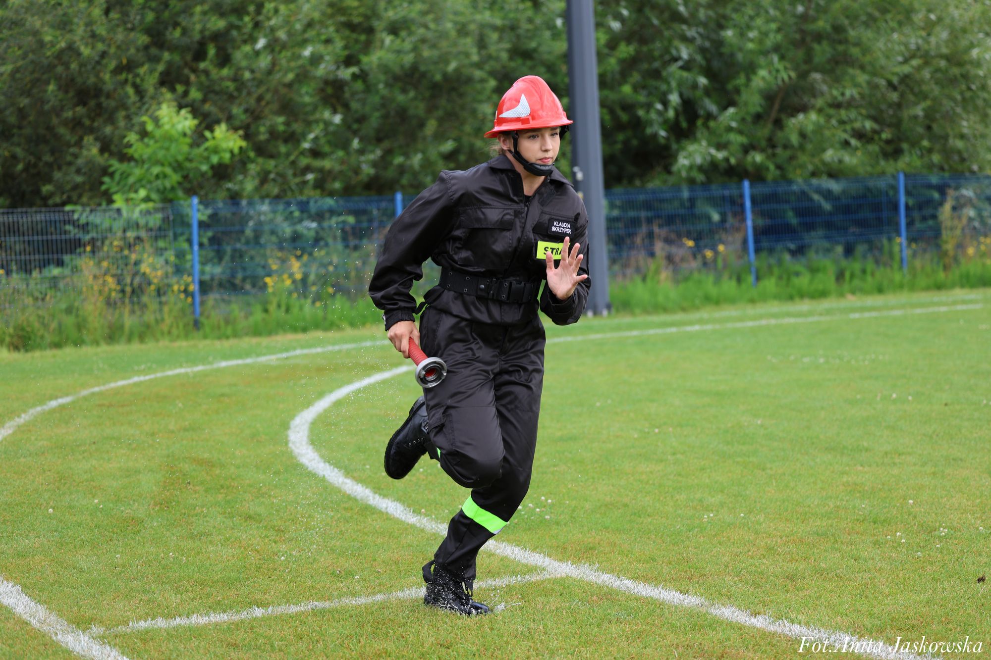 Kobieta w czarnym kombinezonie z odblaskowym paskiem, czerwonym kaskiem i butami biegająca po mokrej trawie na boisku sportowym, trzymająca czerwoną rurę.