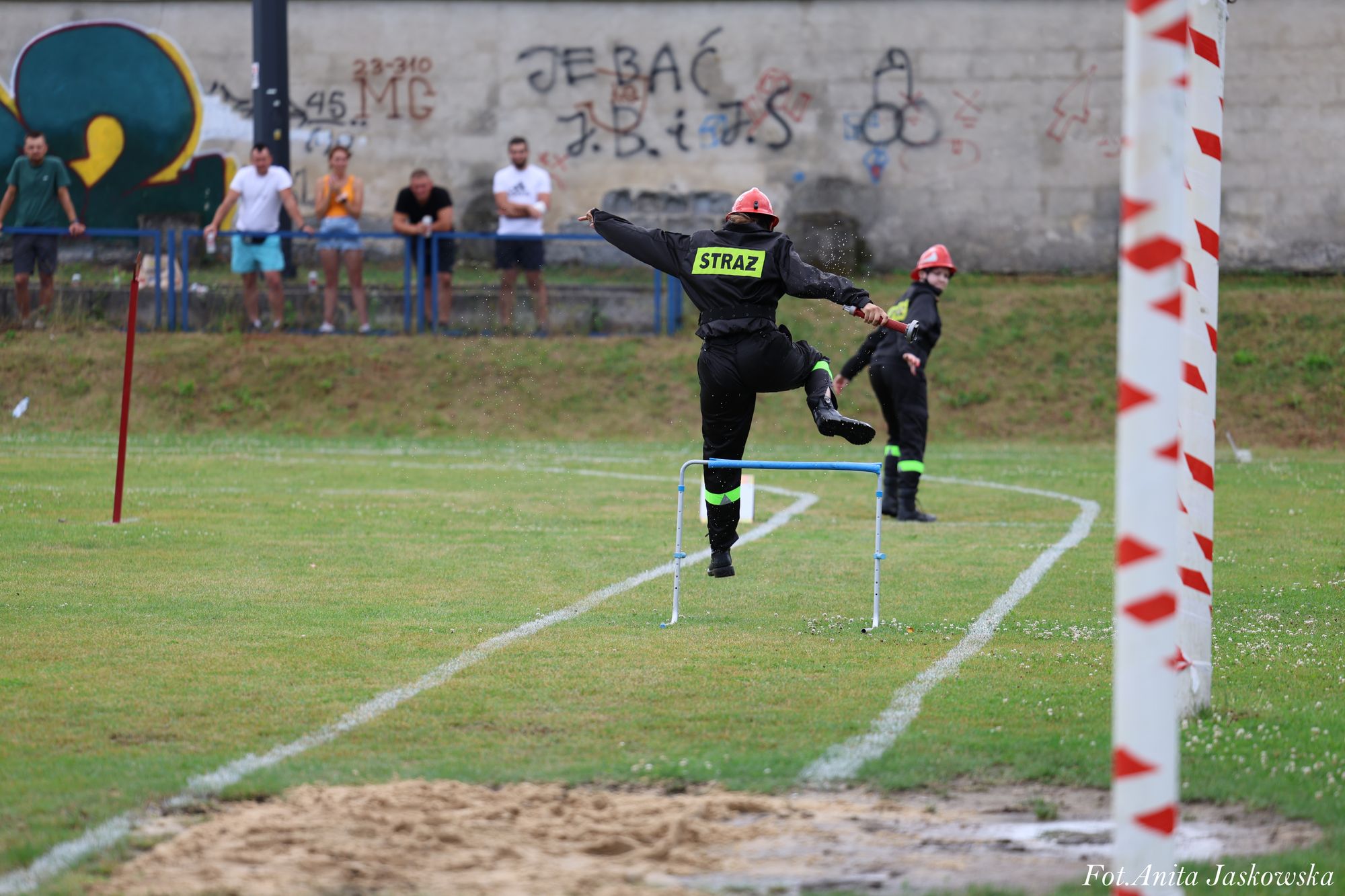 Strażak w czarnym ubraniu i czerwonym hełmie skacze przez płotek na zielonym boisku, w tle mur z graffiti i stojący ludzie.