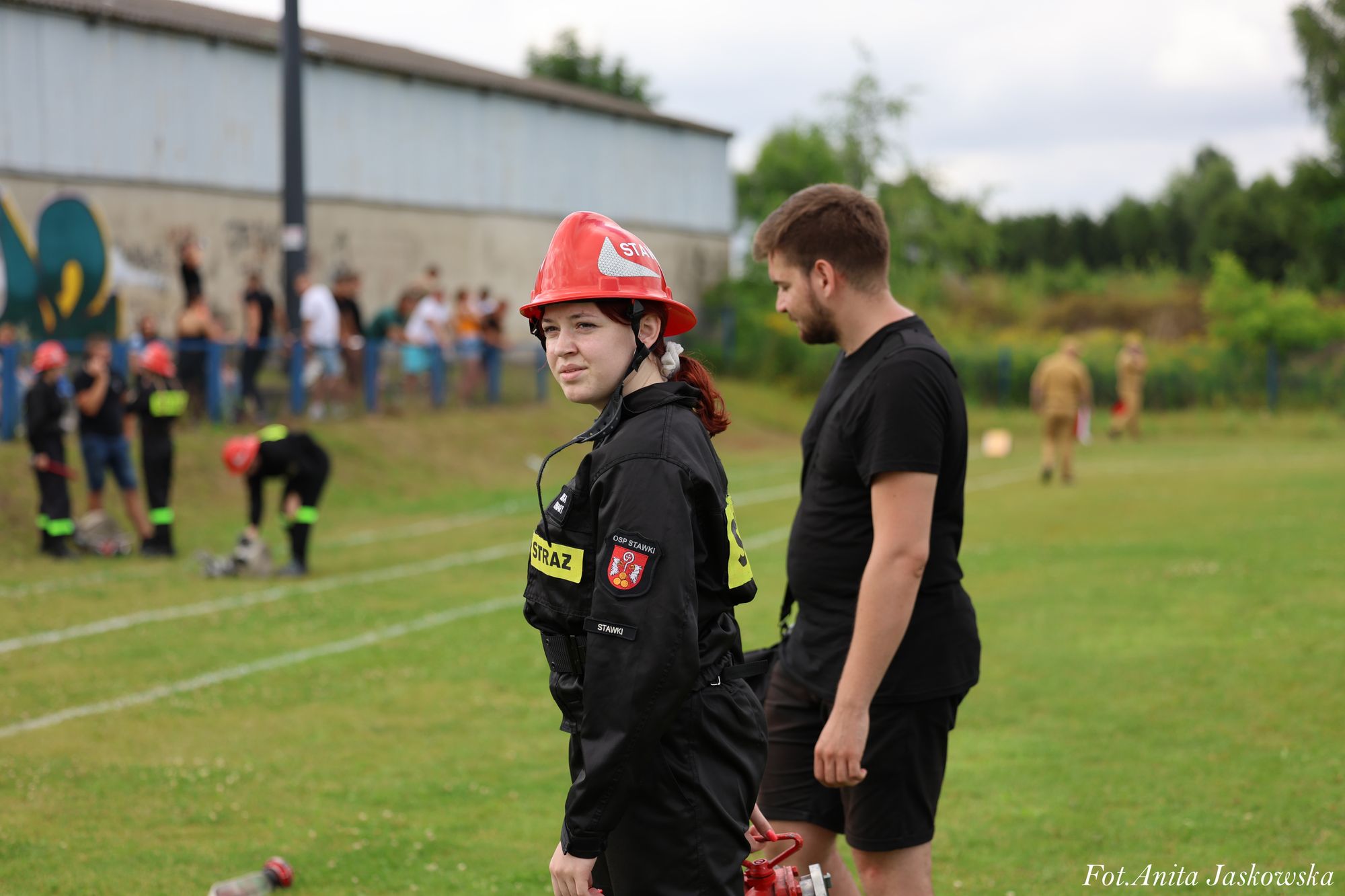 Kobieta w czarnym ubraniu strażackim i czerwonym kasku stoi na zielonym boisku, obok niej mężczyzna w czarnym stroju, w tle ludzie i mur z graffiti.
