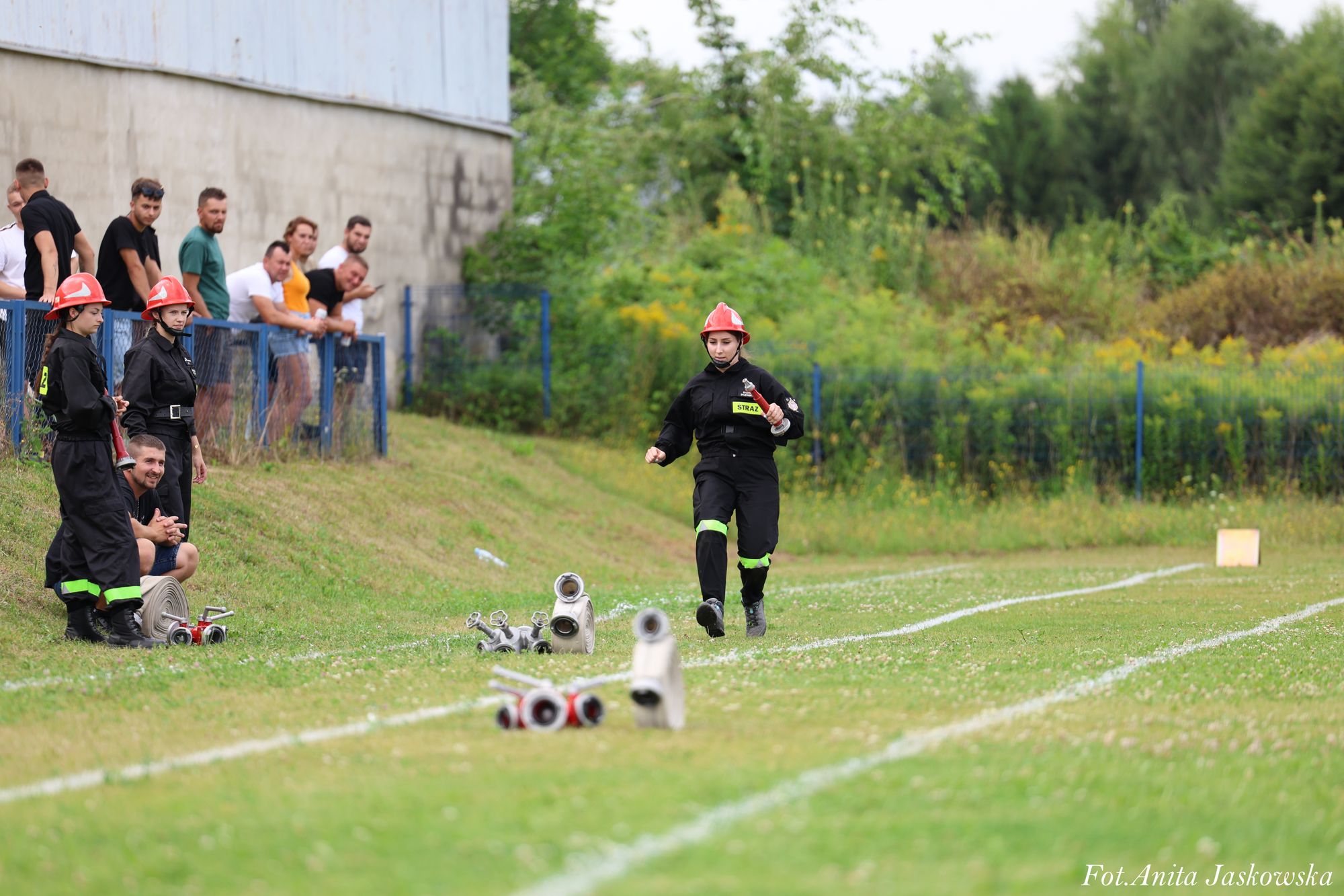 Kobieta w czarnym mundurze strażackim z czerwonym hełmem biegnie po zielonym boisku z rozłożonymi rurami strażackimi, w tle grupa osób obserwujących.