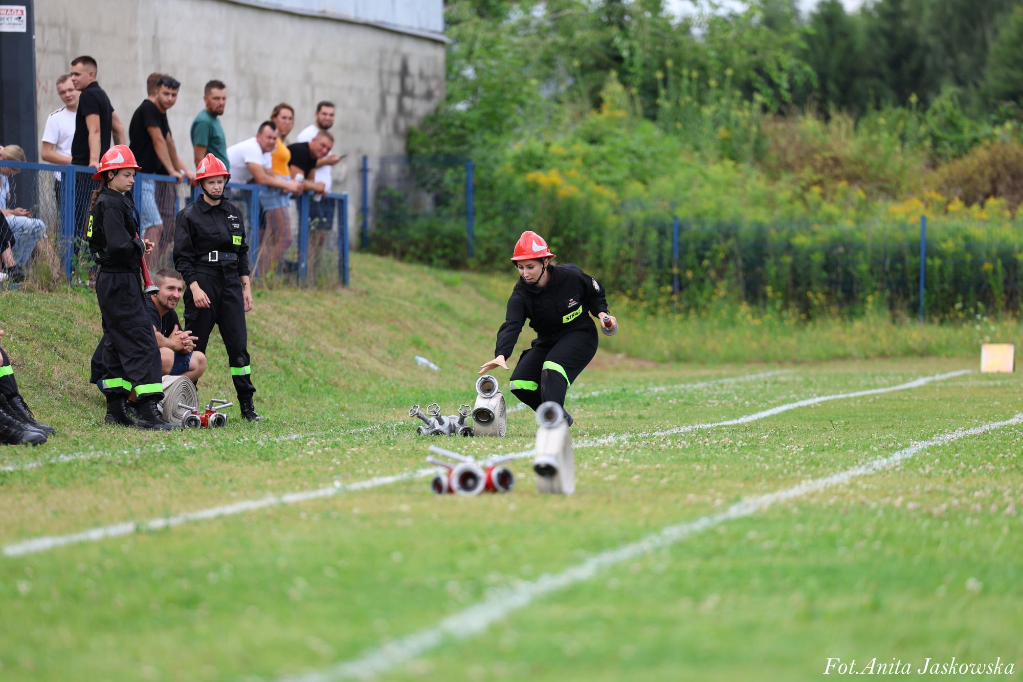 Kobieta w czarnym ubraniu i czerwonym hełmie biegnie po trawiastym boisku z białymi liniami, sięga po zwinięty wąż strażacki, w tle ludzie przy ogrodzeniu i zarośla.