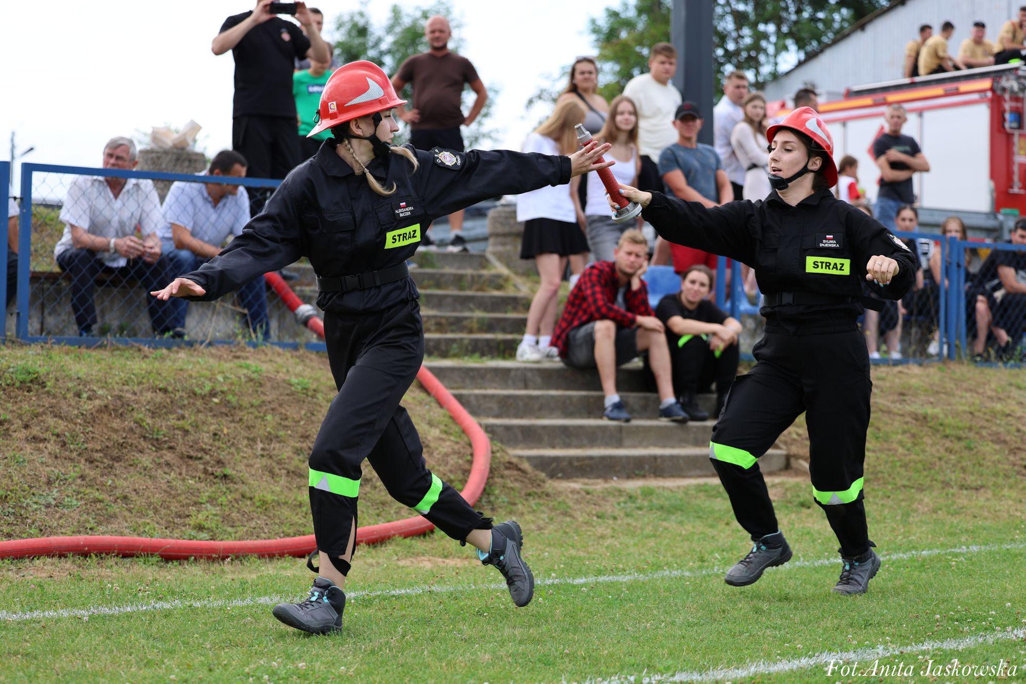 Dwie kobiety w czarnych mundurach strażackich z czerwonymi hełmami przekazują sobie sprzęt na zielonym boisku, w tle grupa stojących i siedzących ludzi oraz wóz strażacki.
