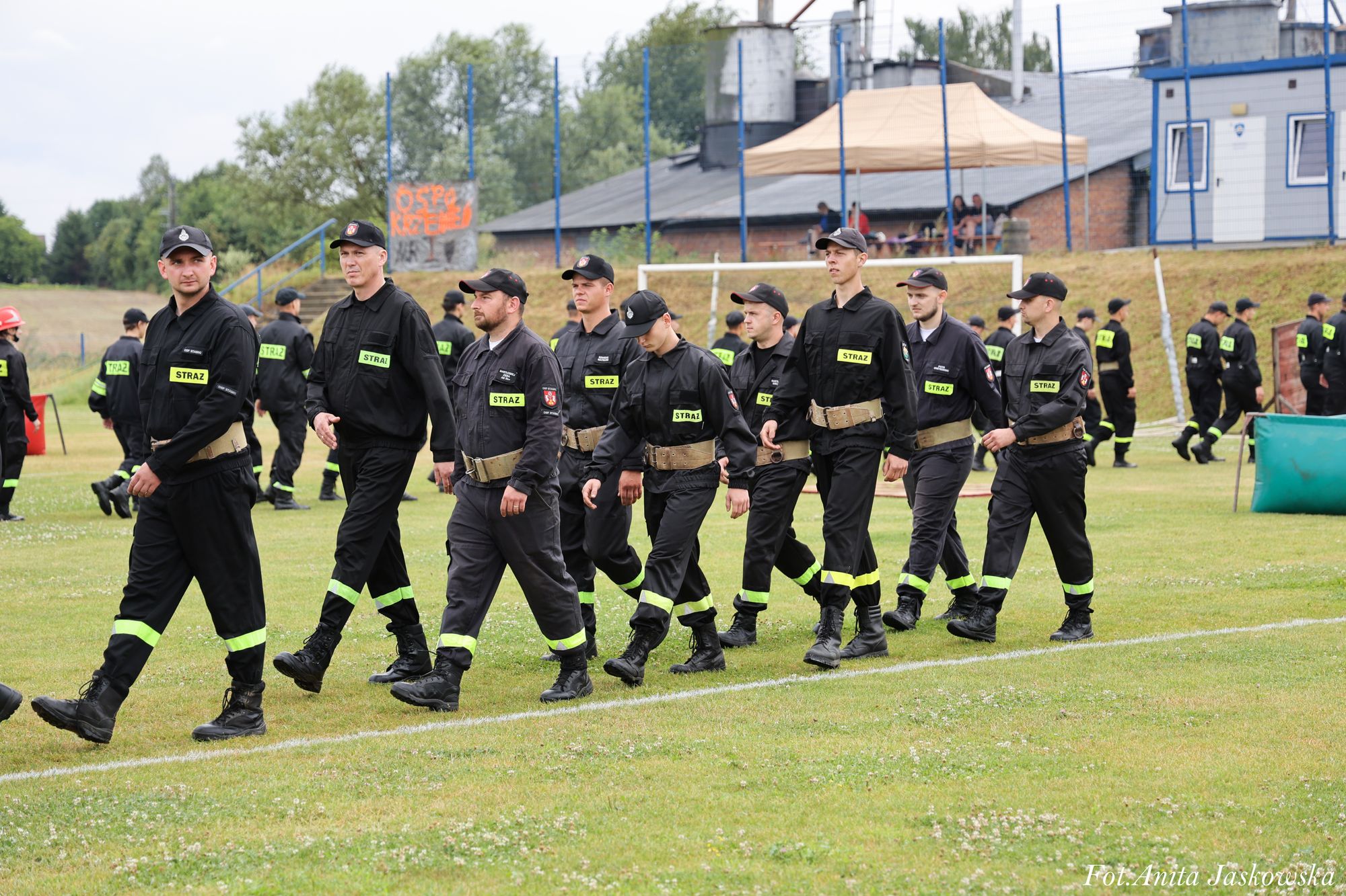 Grupa mężczyzn ubranych w czarne mundury z żółtymi napisami "STRAŻ" maszeruje po zielonym boisku, w tle budynki i ogrodzenie z niebieskimi słupkami.