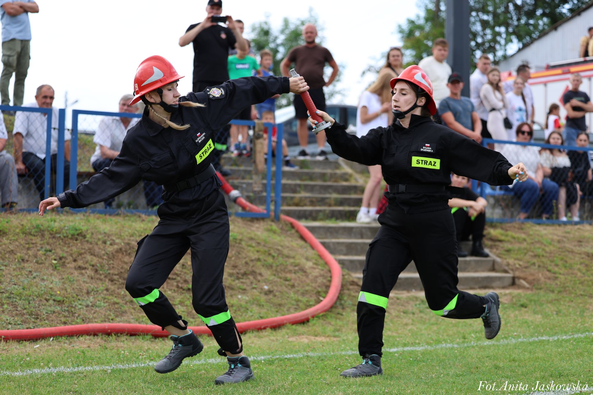 Dwie kobiety w czarnych mundurach strażackich z czerwonymi hełmami przekazują sobie czerwoną rurę na zielonej trawie, w tle ludzie i schody.