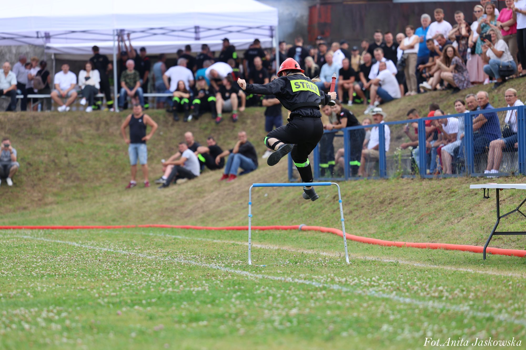 Strażak w czarnym ubraniu i czerwonym hełmie skacze przez przeszkodę na zielonym boisku, w tle tłum widzów i biały namiot.