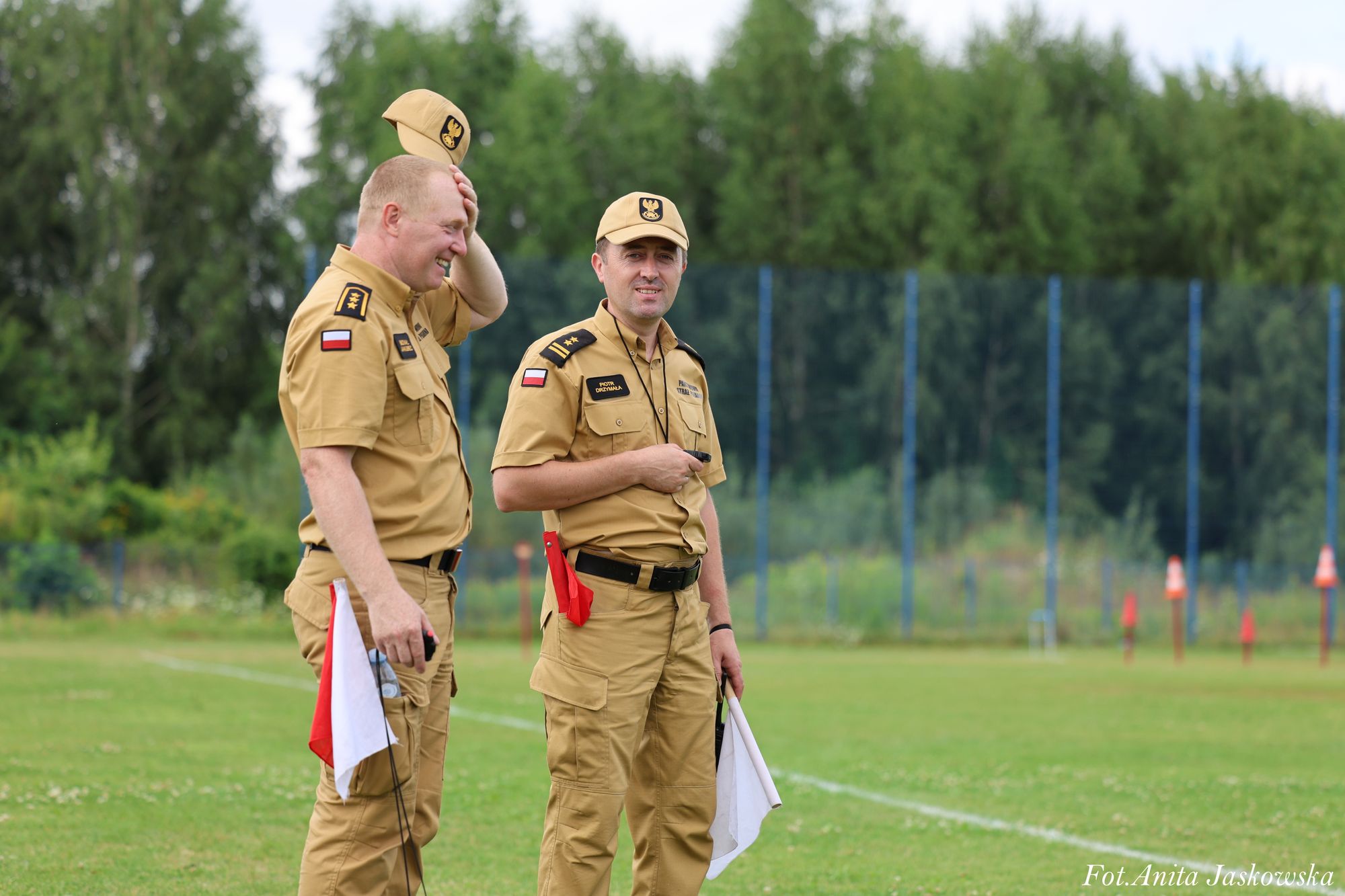 Dwóch mężczyzn w beżowych mundurach z polskimi flagami na rękawach, trzymających biało-czerwone chorągiewki, stojących na zielonym boisku sportowym.