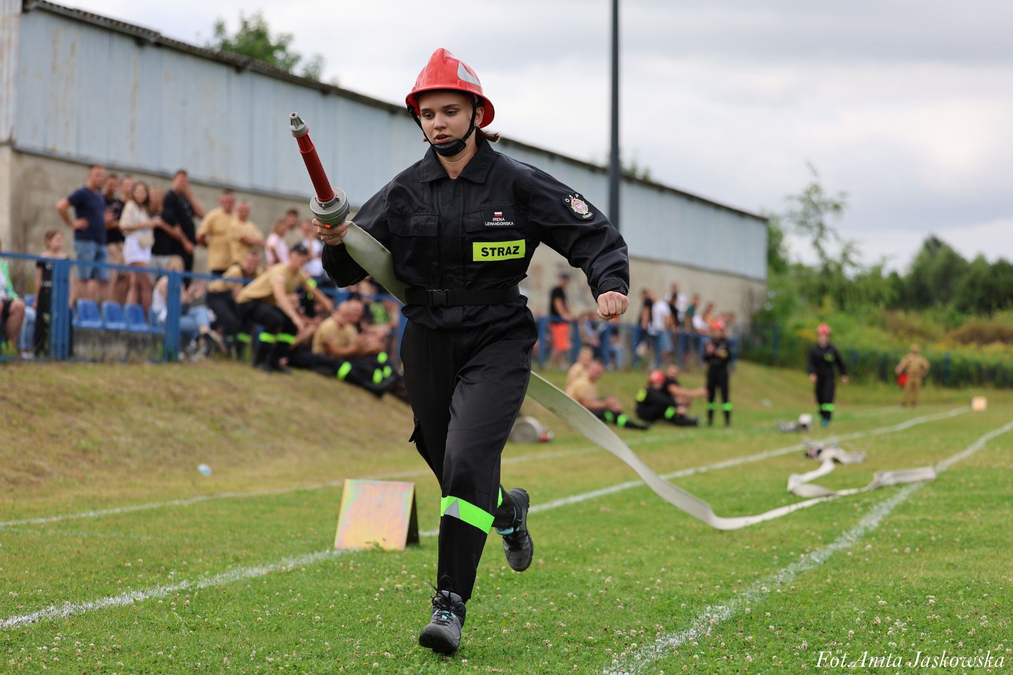 Kobieta w czarnym ubraniu strażackim i czerwonym kasku biegnie po trawie trzymając zwinięty wąż strażacki, w tle rozmyte postacie ludzi i metalowy płot.