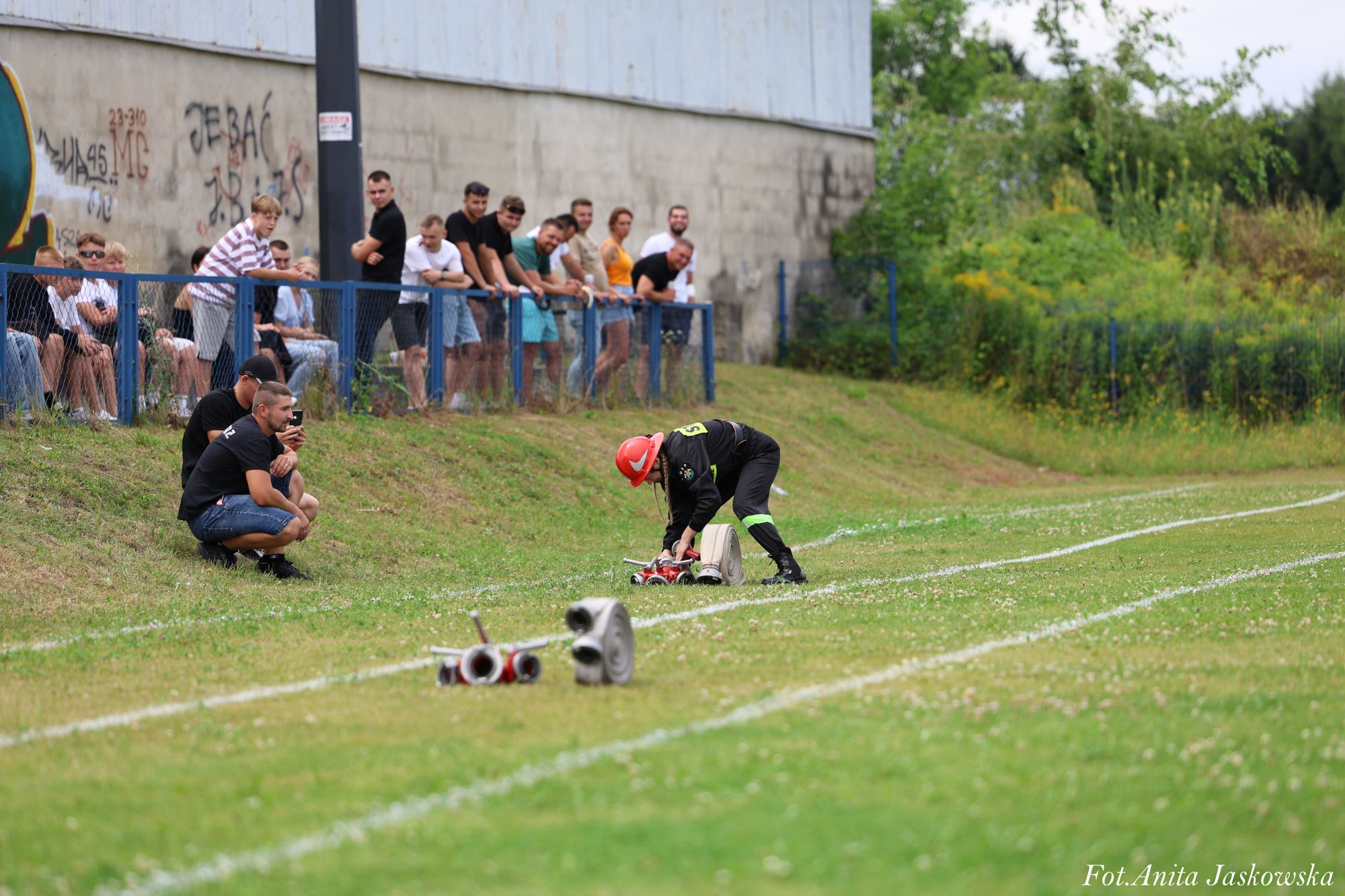 Strażak w czarnym ubraniu i czerwonym hełmie rozkłada sprzęt na trawie, w tle grupa osób przy niebieskim ogrodzeniu i mur z graffiti.
