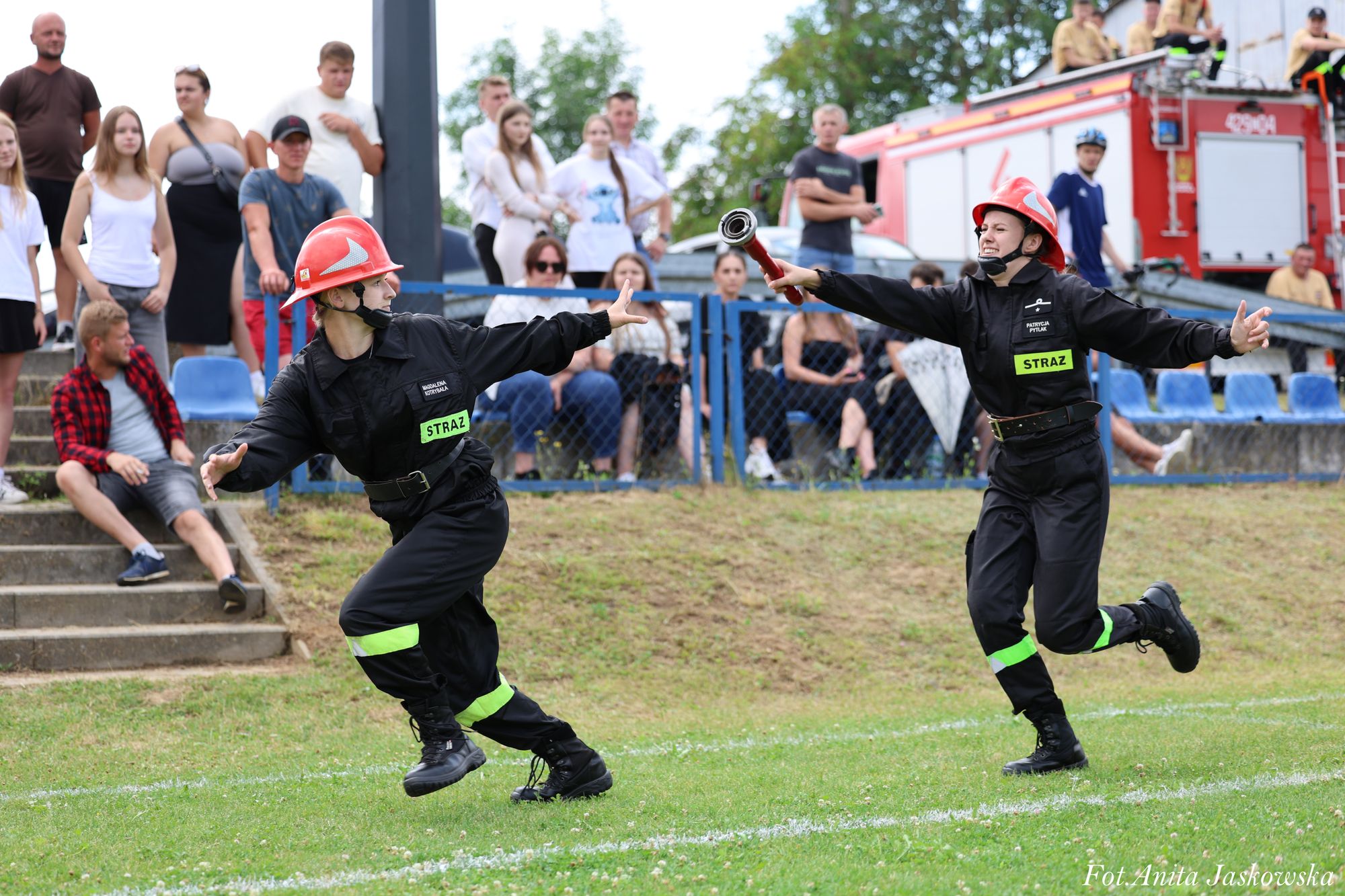 Dwie kobiety w czarnych mundurach strażackich i czerwonych hełmach podczas przekazywania węża strażackiego na zielonym trawniku, w tle widownia i wóz strażacki.