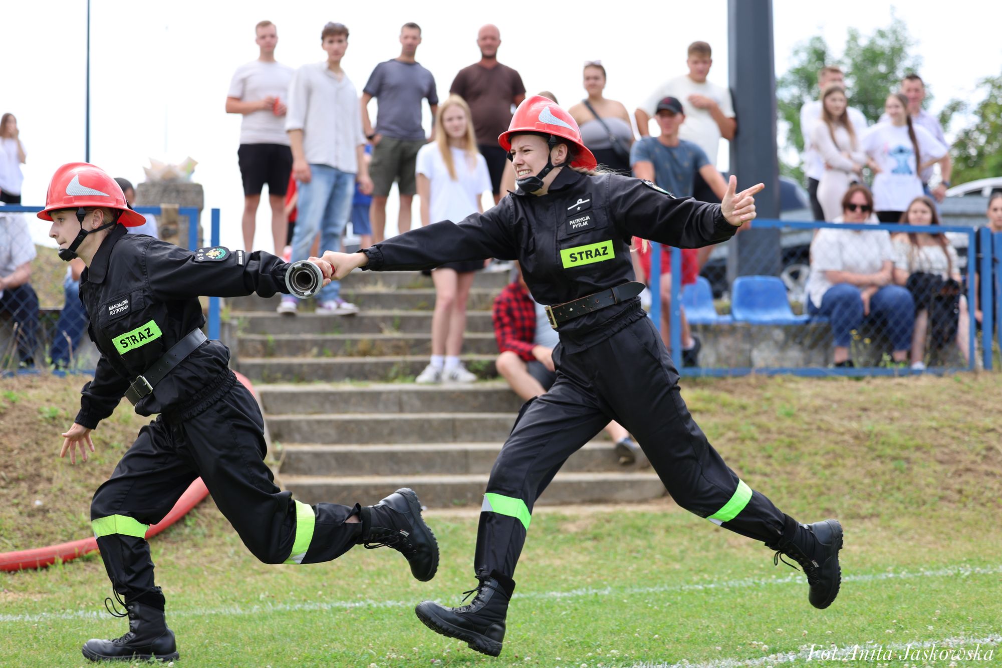Dwie osoby w czarnych mundurach strażackich z czerwonymi hełmami biegną po trawie, trzymając się za ręce, w tle stoi grupa osób na schodach i na trawie.