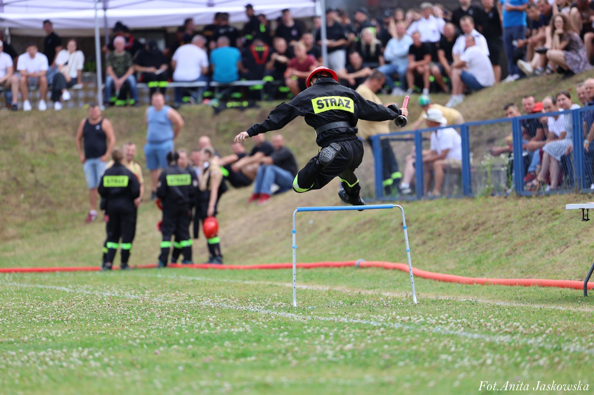 Strażak w czarnym ubraniu i czerwonym hełmie skacze przez niebieską przeszkodę na zielonym boisku, w tle widoczna grupa ludzi i widzowie za niebieską barierką.