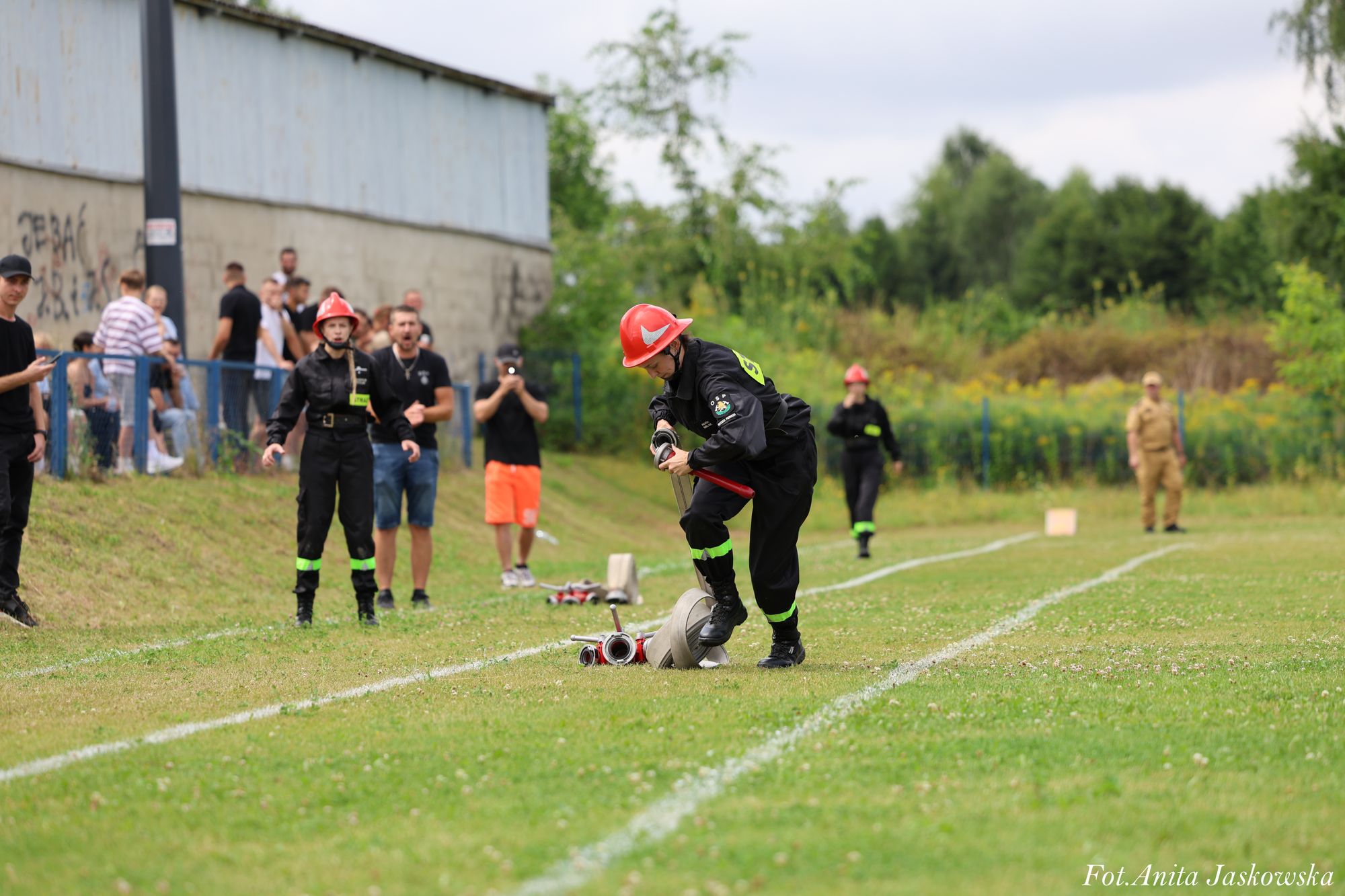 Strażak w czerwonym kasku i czarnym mundurze z zielonymi odblaskami rozkłada wąż strażacki na trawiastym boisku, w tle ludzie i budynek z szarą ścianą.