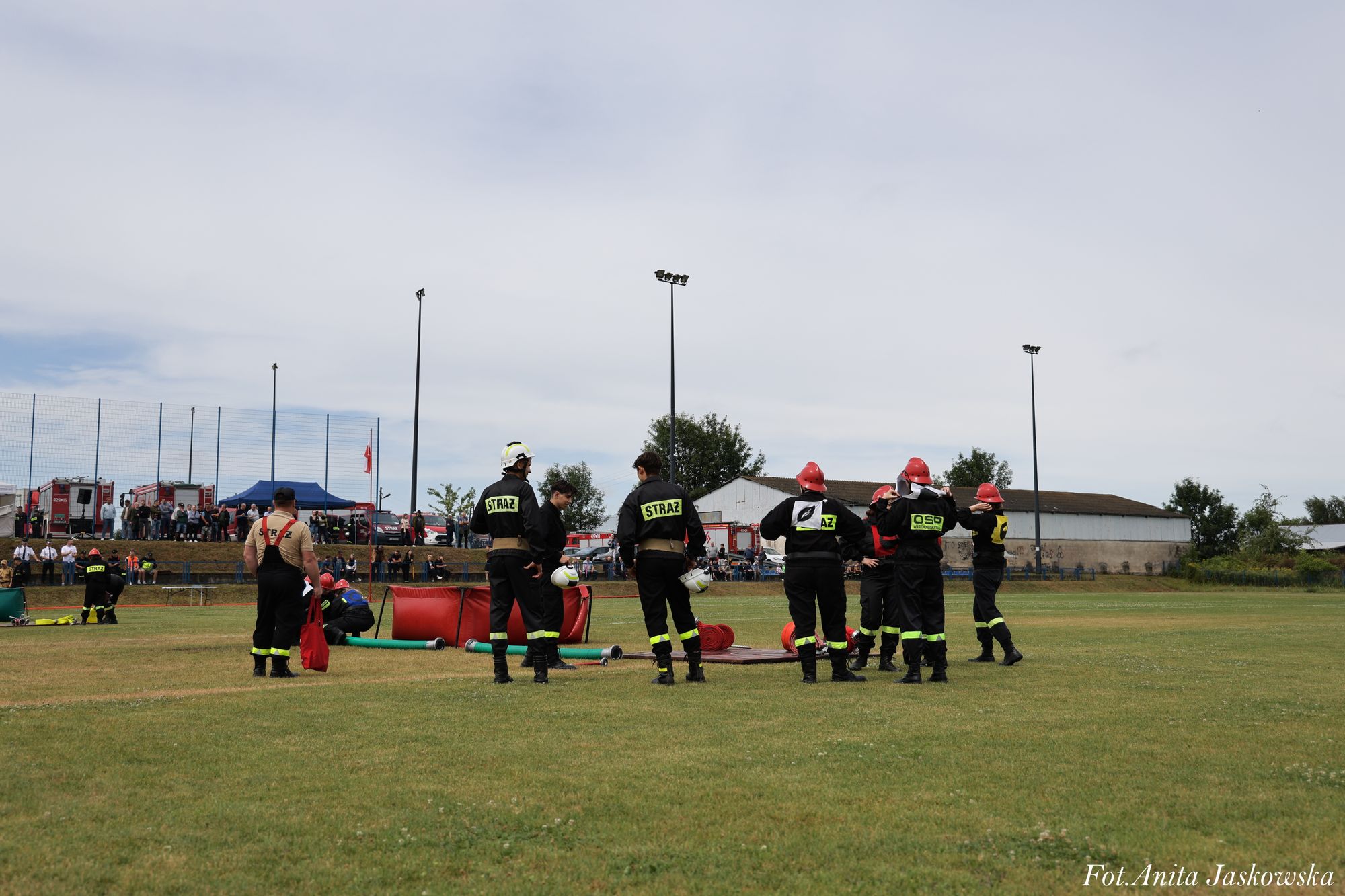 Grupa strażaków w czarnych mundurach z żółtymi napisami stoi na zielonym boisku, w tle ludzie, wozy strażackie i budynki.