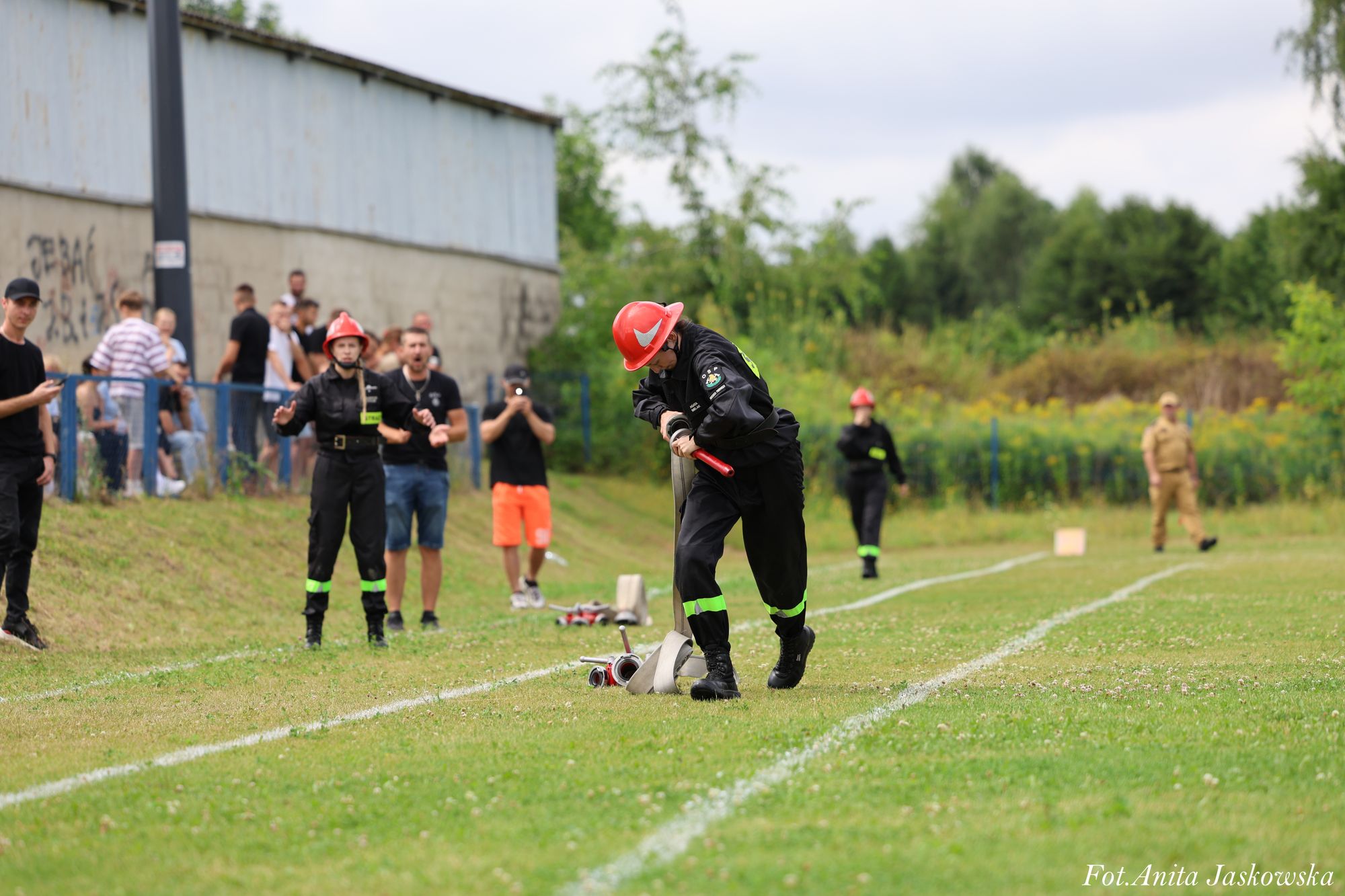 Osoba w czarnym ubraniu strażaka i czerwonym hełmie zwija beżowy wąż na zielonym boisku z białymi liniami, w tle inni ludzie i zarośla.