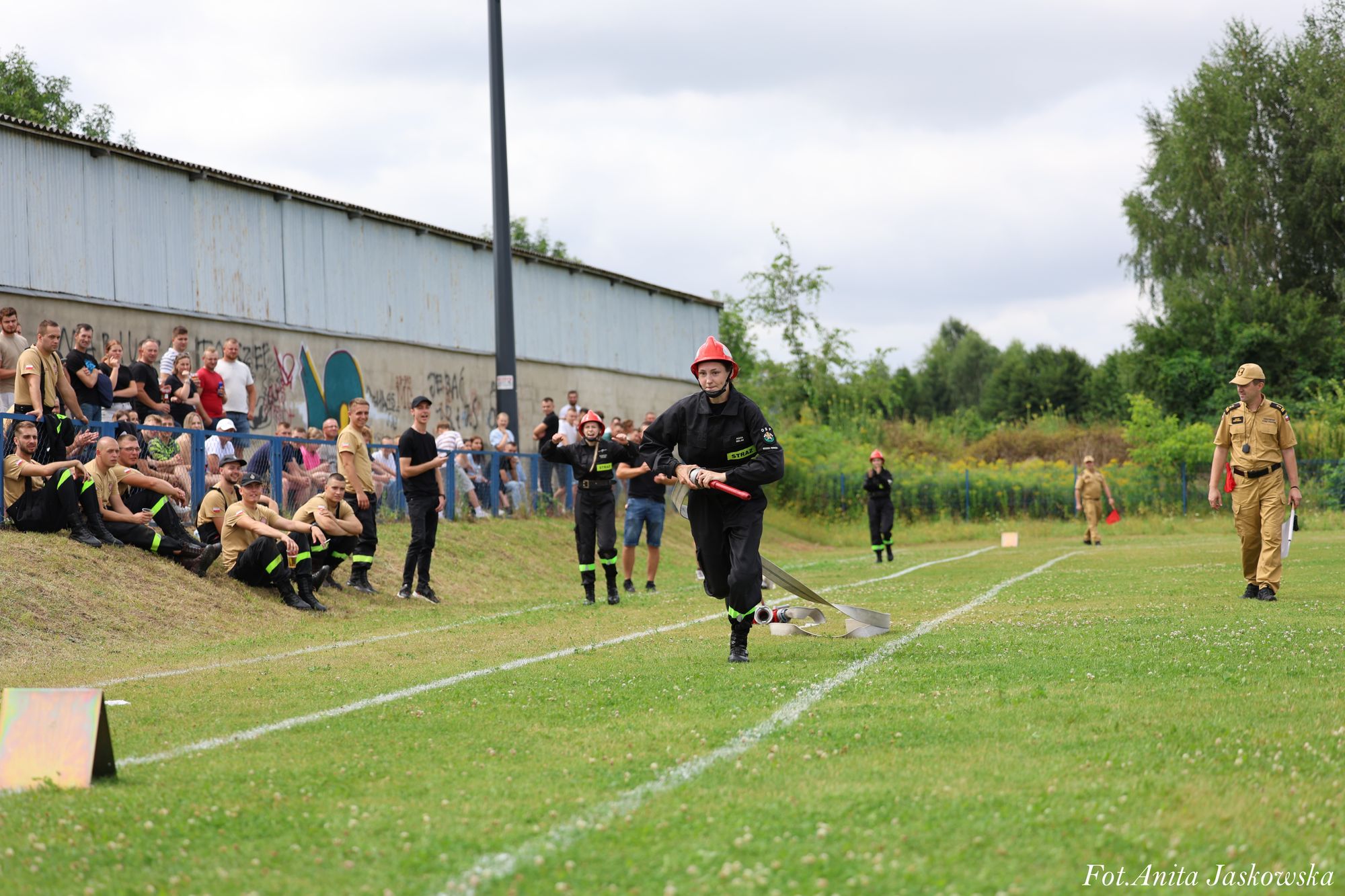 Młoda strażaczka w czarnym mundurze i czerwonym hełmie biegnie po trawiastej bieżni z rozwiniętym wężem strażackim, obok sędzia i widzowie na tle budynku i zieleni.