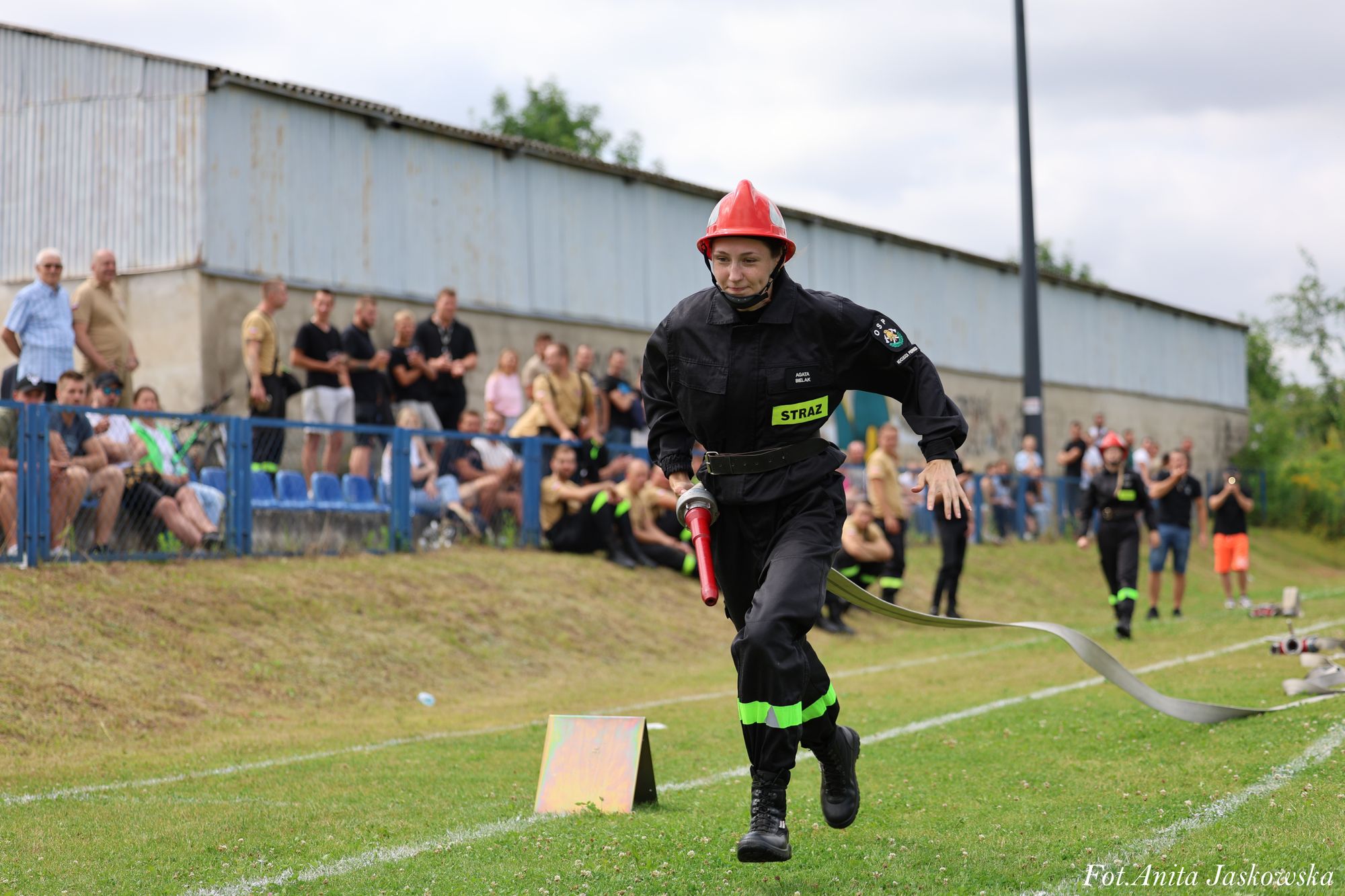 Strażaczka w czarnym ubraniu ochronnym i czerwonym hełmie biegnie po zielonym boisku z rozwiniętym wężem strażackim, w tle ludzie na trybunach i inni strażacy.