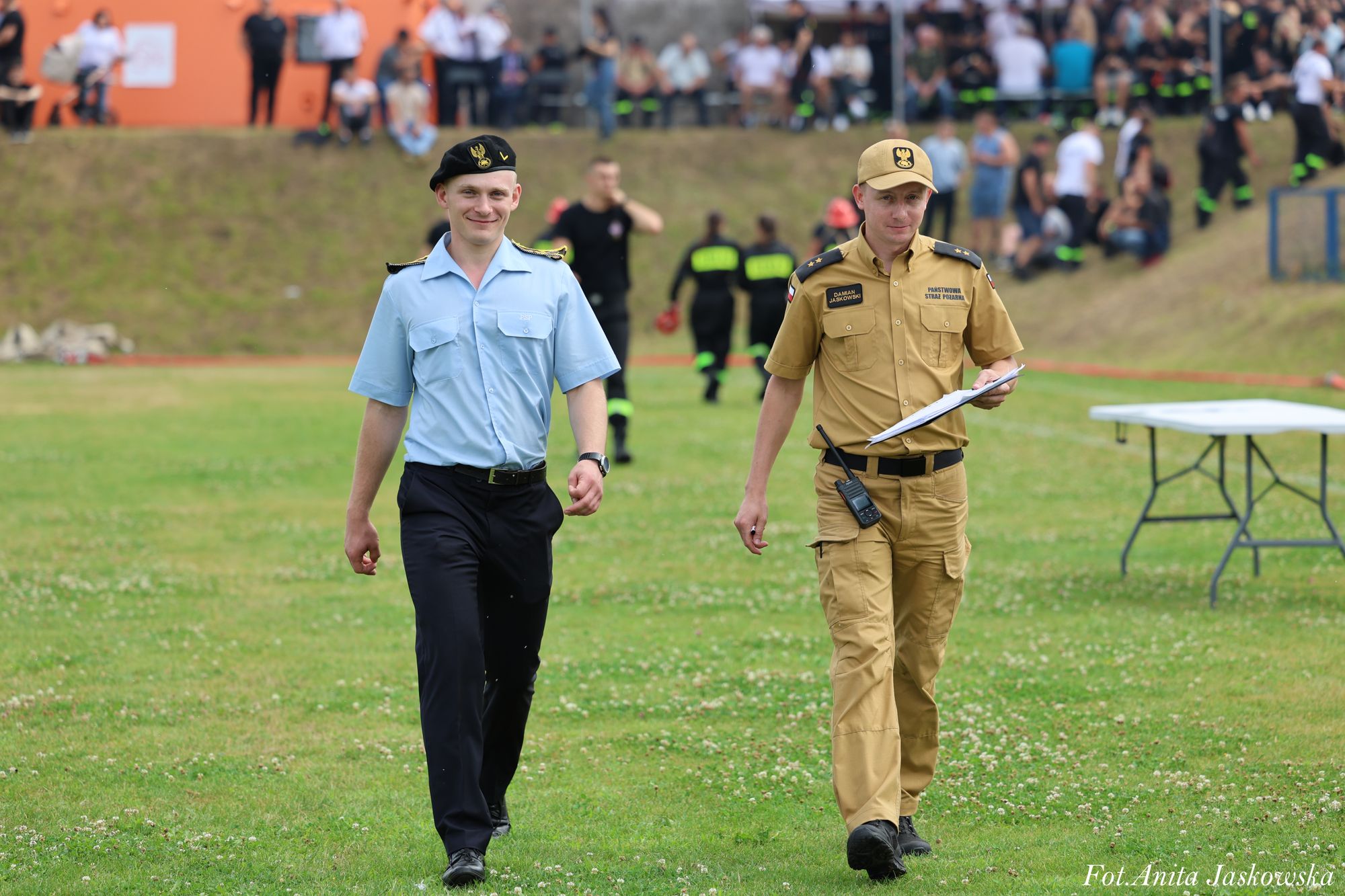 Dwóch mężczyzn w mundurach idzie po trawie, jeden w niebieskiej koszuli i czarnym berecie, drugi w beżowym mundurze i czapce, trzyma dokumenty i radiotelefon.