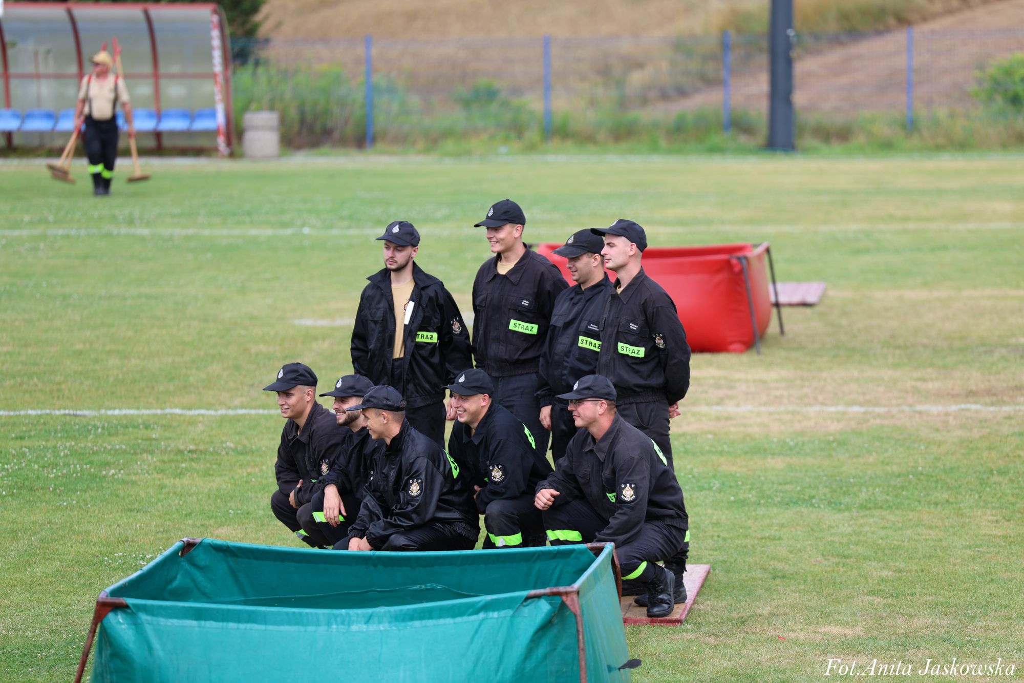 Dziesięciu mężczyzn w czarnych mundurach i czapkach z napisem "STRAŻ" na zielonych łatach, pozujących na trawie, przed nimi zielony zbiornik i czerwony zbiornik w tle.