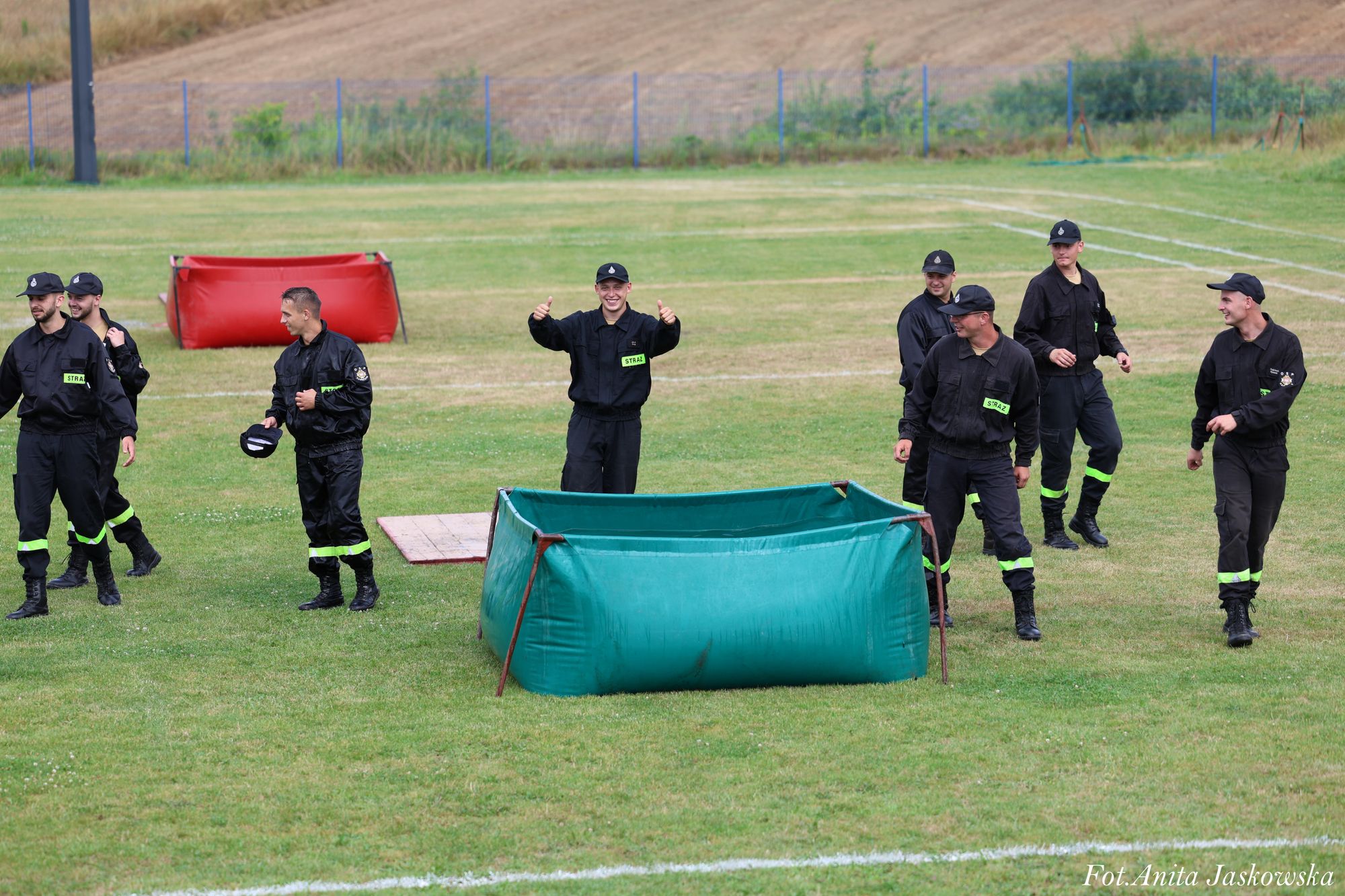 Grupa mężczyzn w czarnych mundurach z zielonymi naszywkami "STRAŻ" stoi na zielonym trawniku obok zielonego i czerwonego pojemnika na metalowych stelażach.