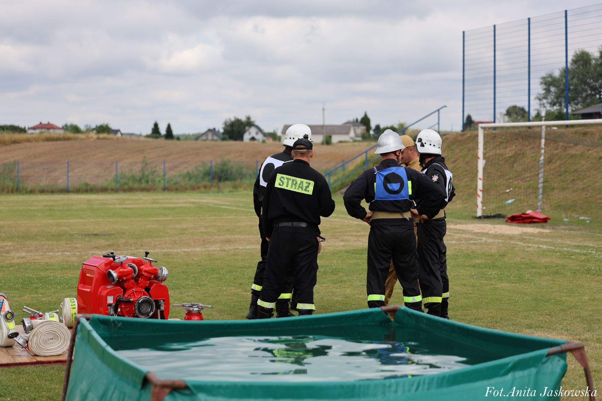 Czterech strażaków w czarnych mundurach z neonowymi elementami i białych hełmach stoi na zielonym boisku sportowym obok czerwonej pompy i złożonych węży strażackich, z przodu zielony zbiornik z wodą.