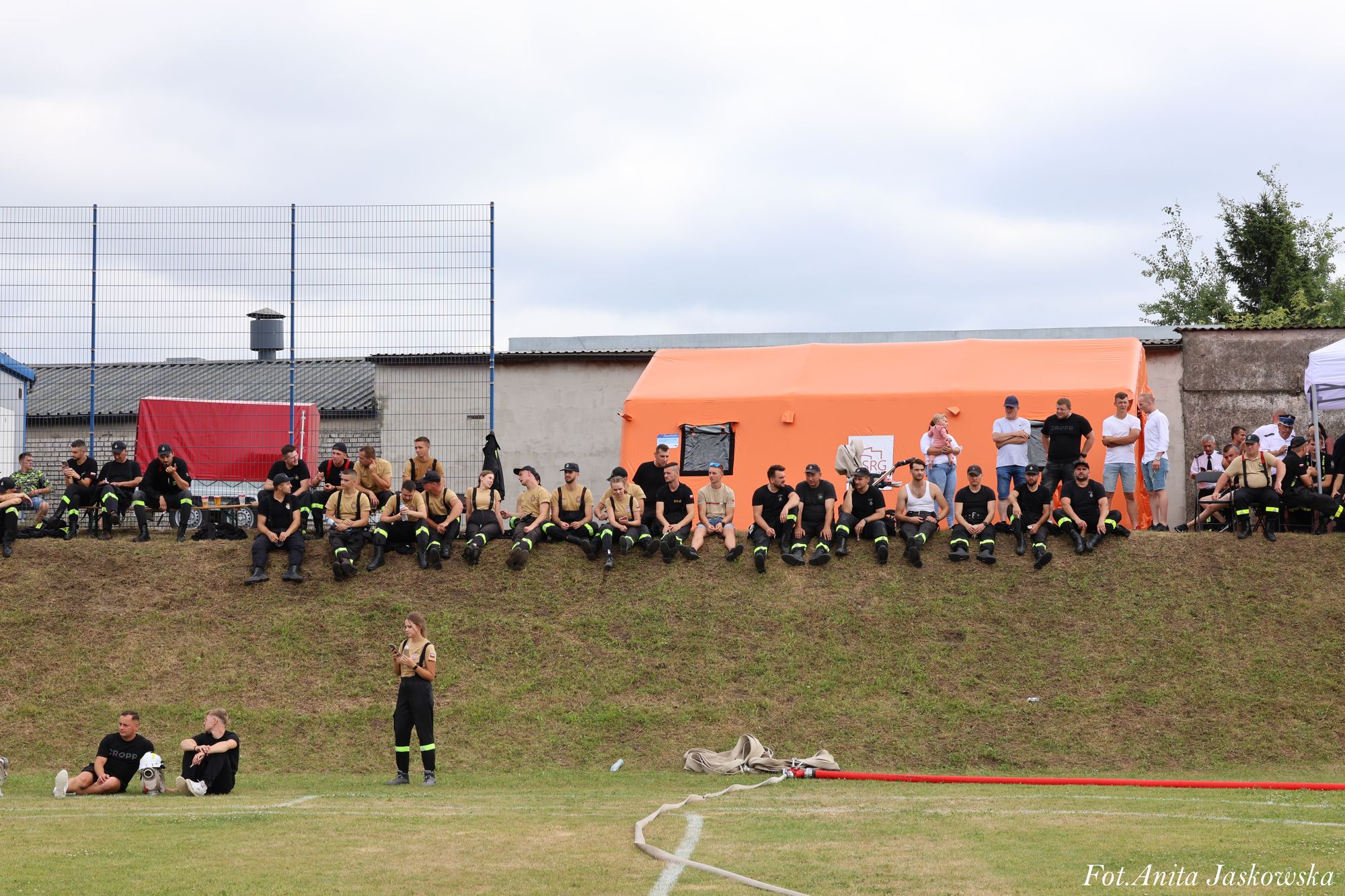 Grupa osób w mundurach strażackich siedzi na skarpie, za nimi pomarańczowy namiot i budynki, na pierwszym planie zielona trawa i rozwinięty wąż strażacki.