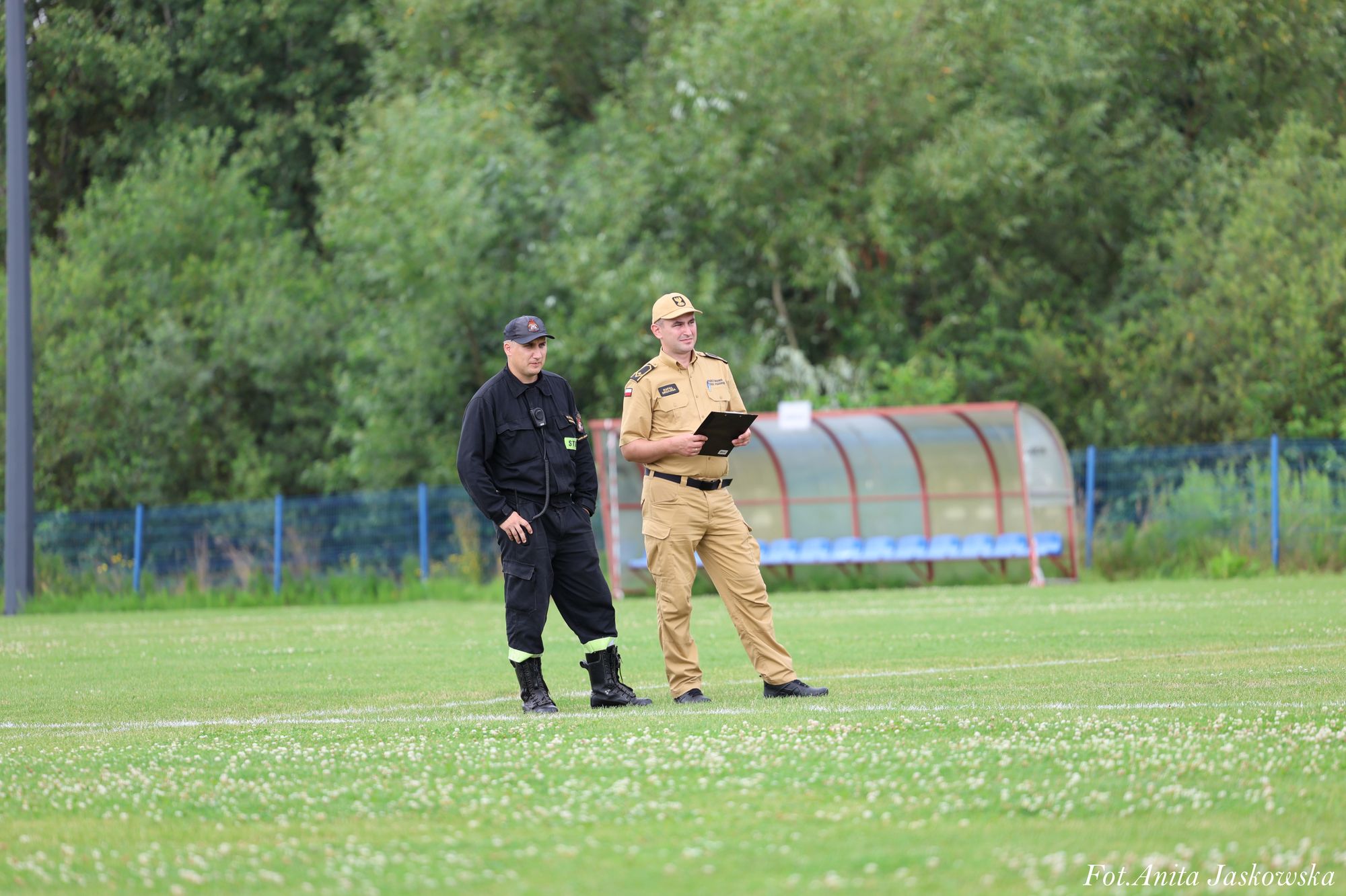Dwaj mężczyźni w mundurach, jeden czarny z napisem "STRAŻ", drugi beżowy z czapką, stoją na zielonym boisku sportowym z niebieskimi ławkami i zalesionym tłem.