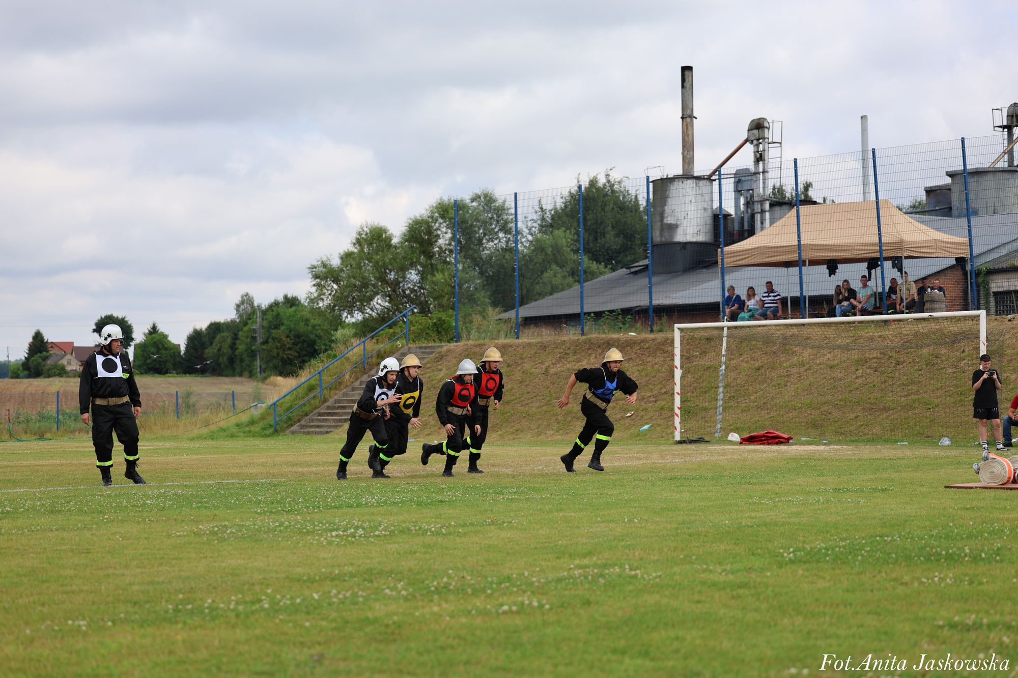 Pięć osób w mundurach strażackich biegnie po zielonym boisku, z tyłu bramka, widzowie pod beżowym namiotem, budynki przemysłowe i drzewa.