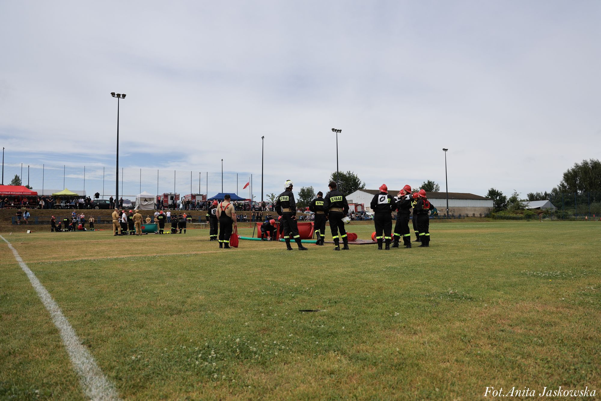 Grupa strażaków w czarnych mundurach i czerwonych hełmach na zielonym boisku sportowym, w tle trybuny z ludźmi i namioty.
