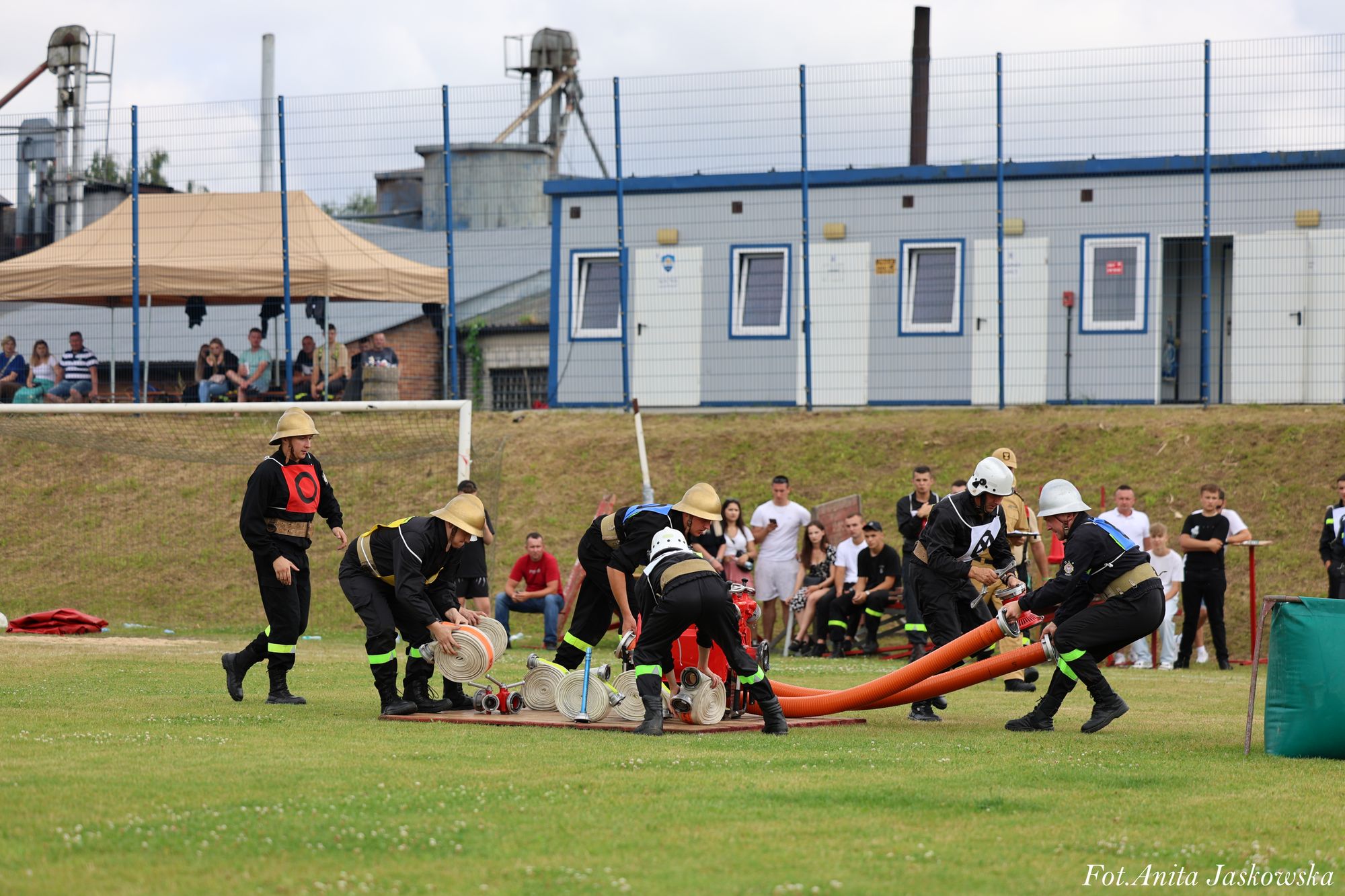 Grupa strażaków w czarnych mundurach i hełmach rozkłada węże strażackie na zielonym boisku, w tle widoczni widzowie i budynki.