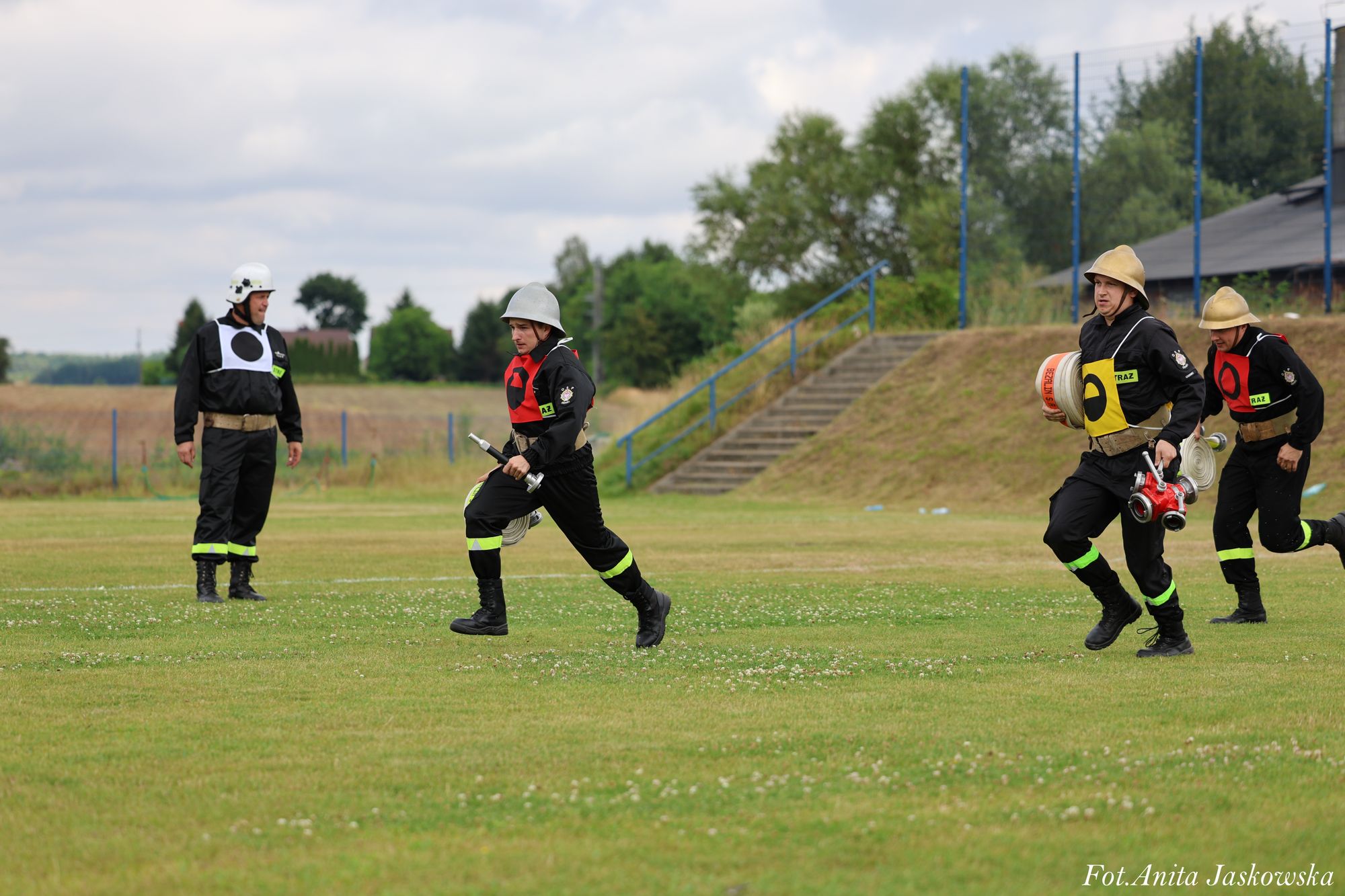 Czterech strażaków w czarnych mundurach i hełmach na zielonej trawie, trzech biegnie, jeden stoi na tle schodów i drzew.