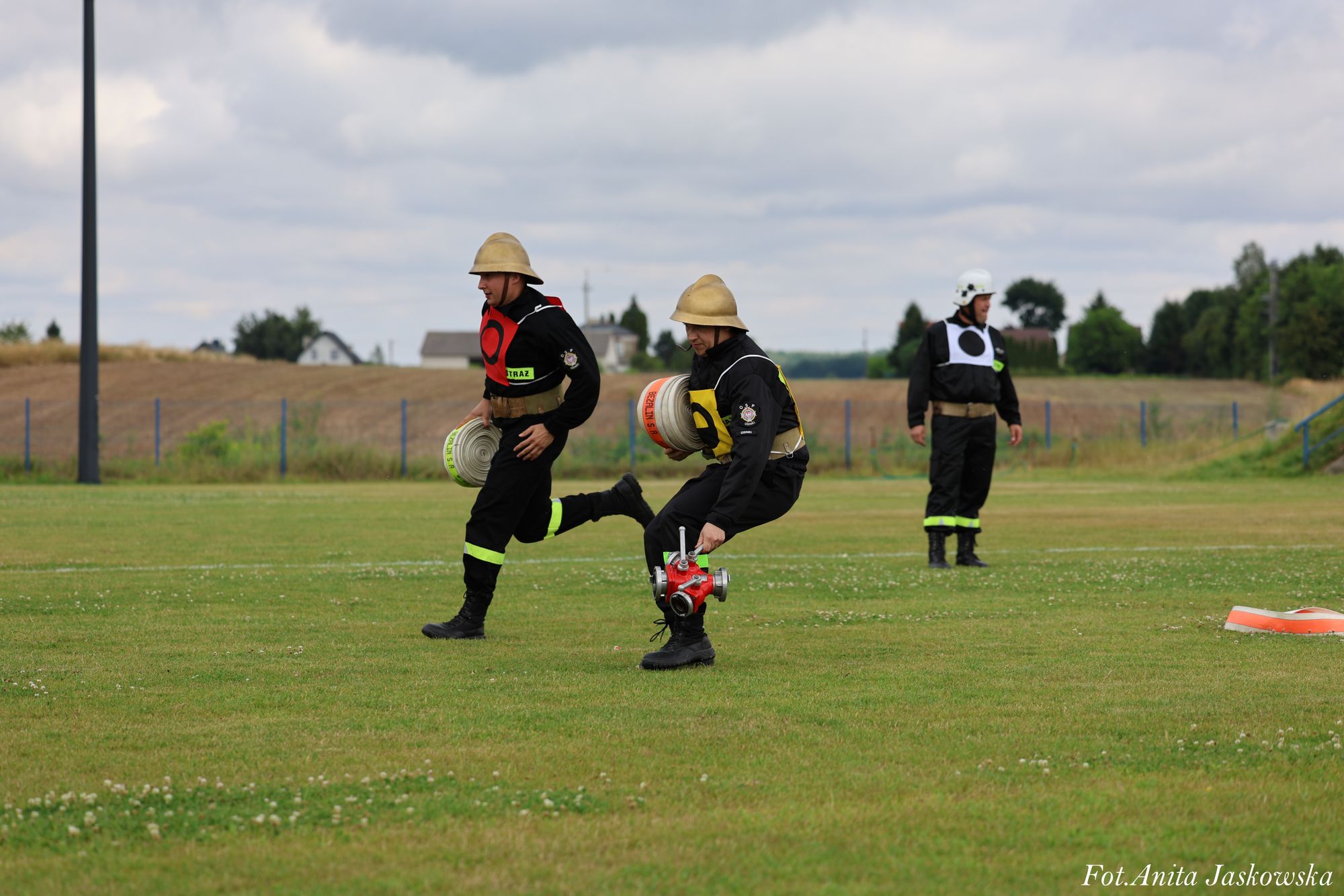 Trzech strażaków na zielonej łące, dwóch w hełmach beżowych, jeden w białym kasku, ubrani w czarne mundury z odblaskowymi paskami, dwóch niesie zwinięte węże strażackie.