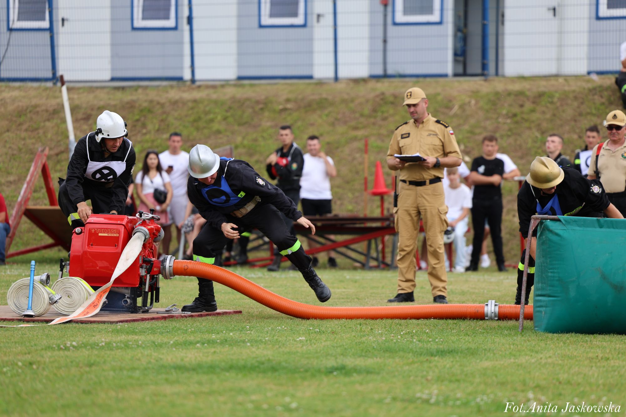 Trzech strażaków w czarnych mundurach i hełmach obsługuje czerwoną pompę z pomarańczowym wężem na zielonej trawie, w tle ludzie i budynek z niebieskimi oknami.