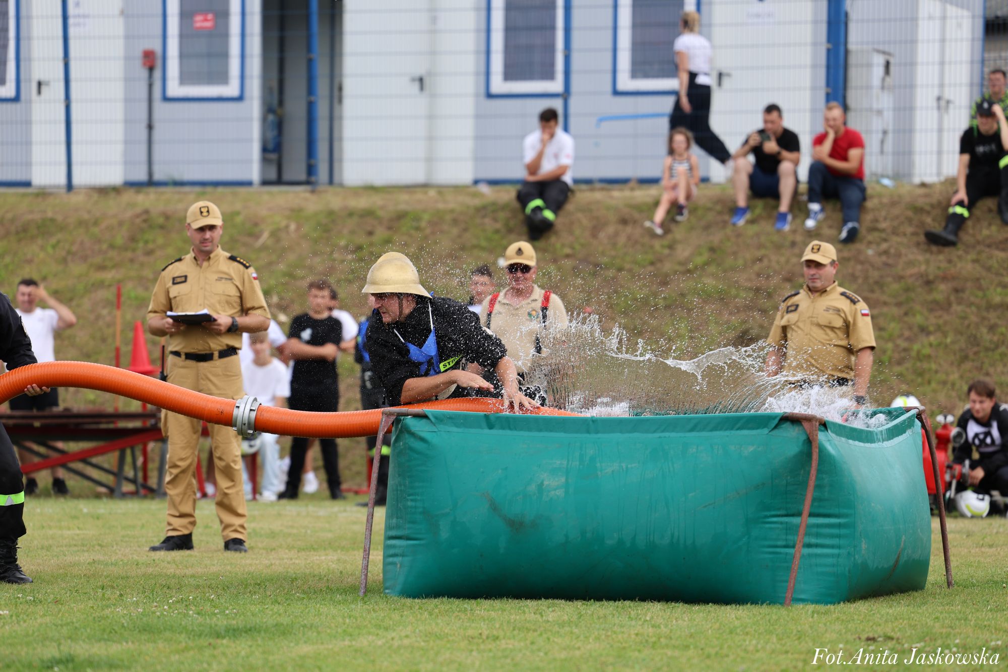 Strażak w helmie wyciąga pomarańczowy wąż z dużego zielonego zbiornika z wodą na trawie, w tle ludzie obserwujący wydarzenie.