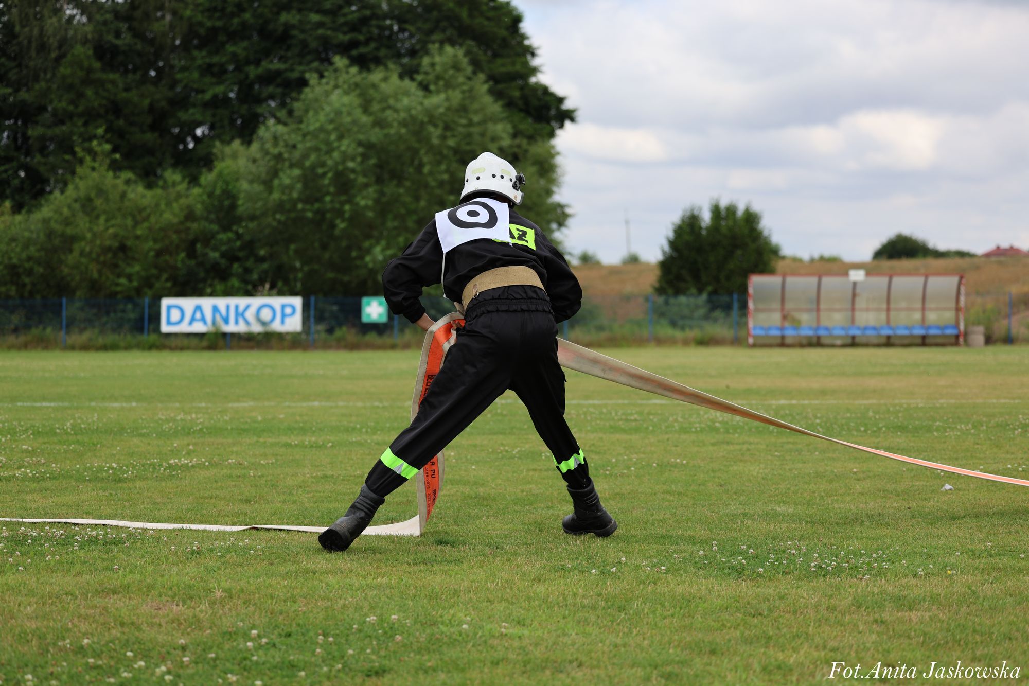 Strażak w czarnym ubraniu z odblaskowymi paskami i białym hełmem rozkłada pomarańczowo-biały wąż strażacki na zielonym boisku sportowym.